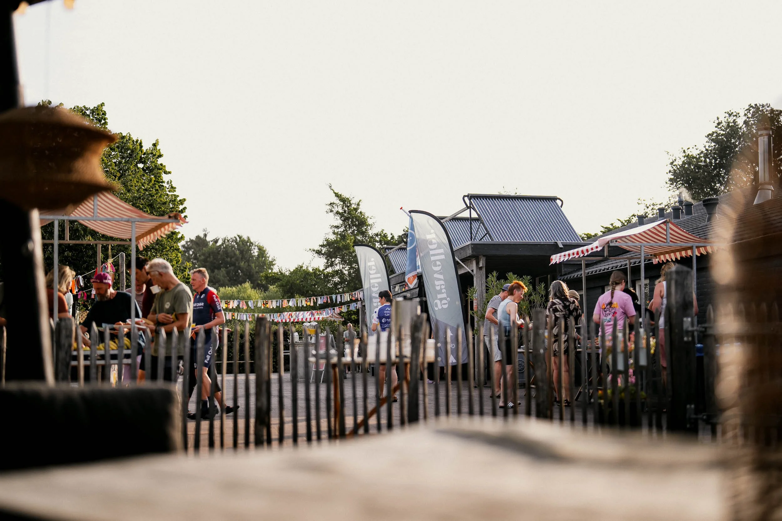 People gathering outdoors at a social event with flags and umbrellas, some standing near tables and engaging in conversations.