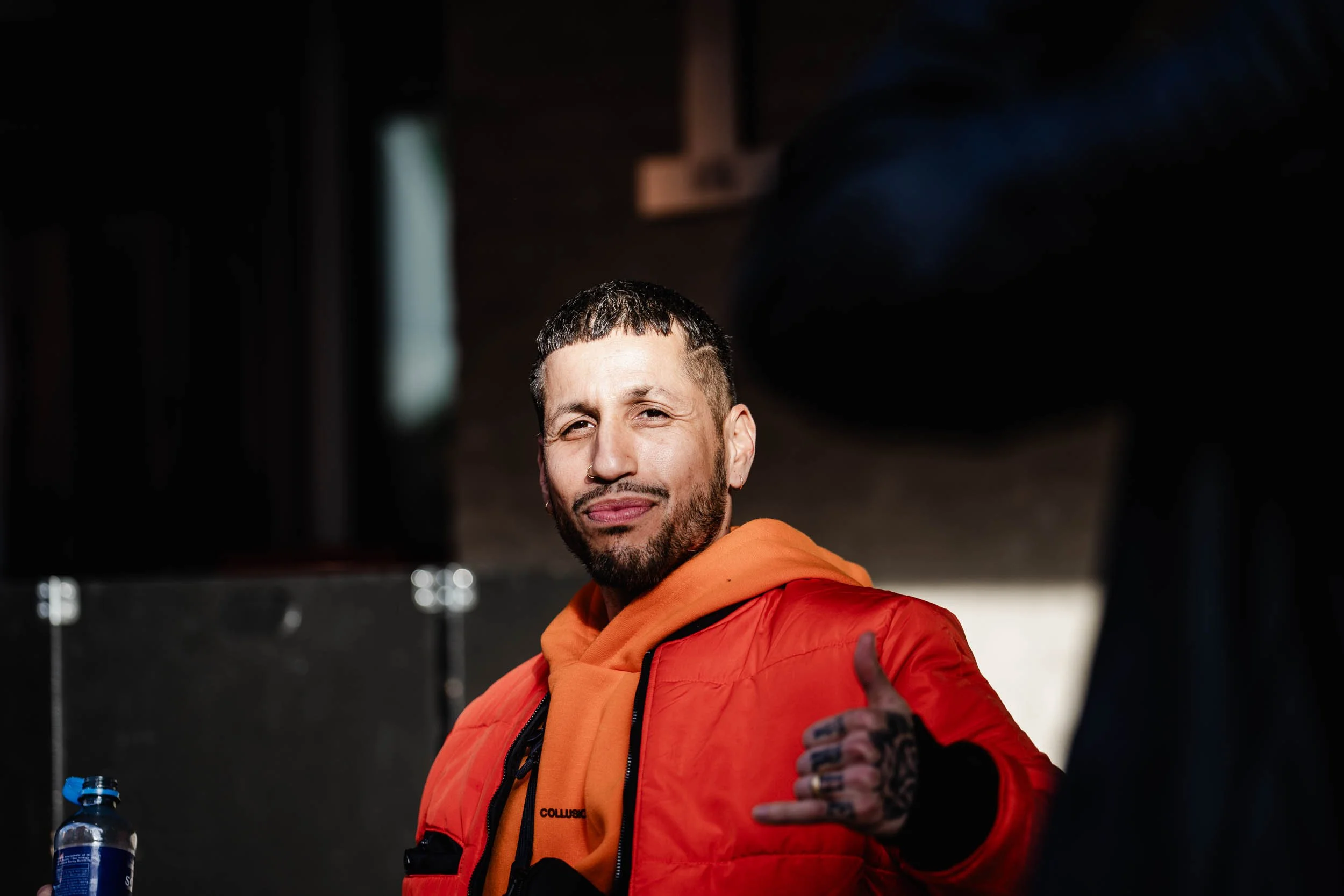 A young man with tattoos on his hand and a beard, wearing an orange hoodie and red jacket, giving a thumbs-up and smiling at the camera in a dimly lit indoor setting.
