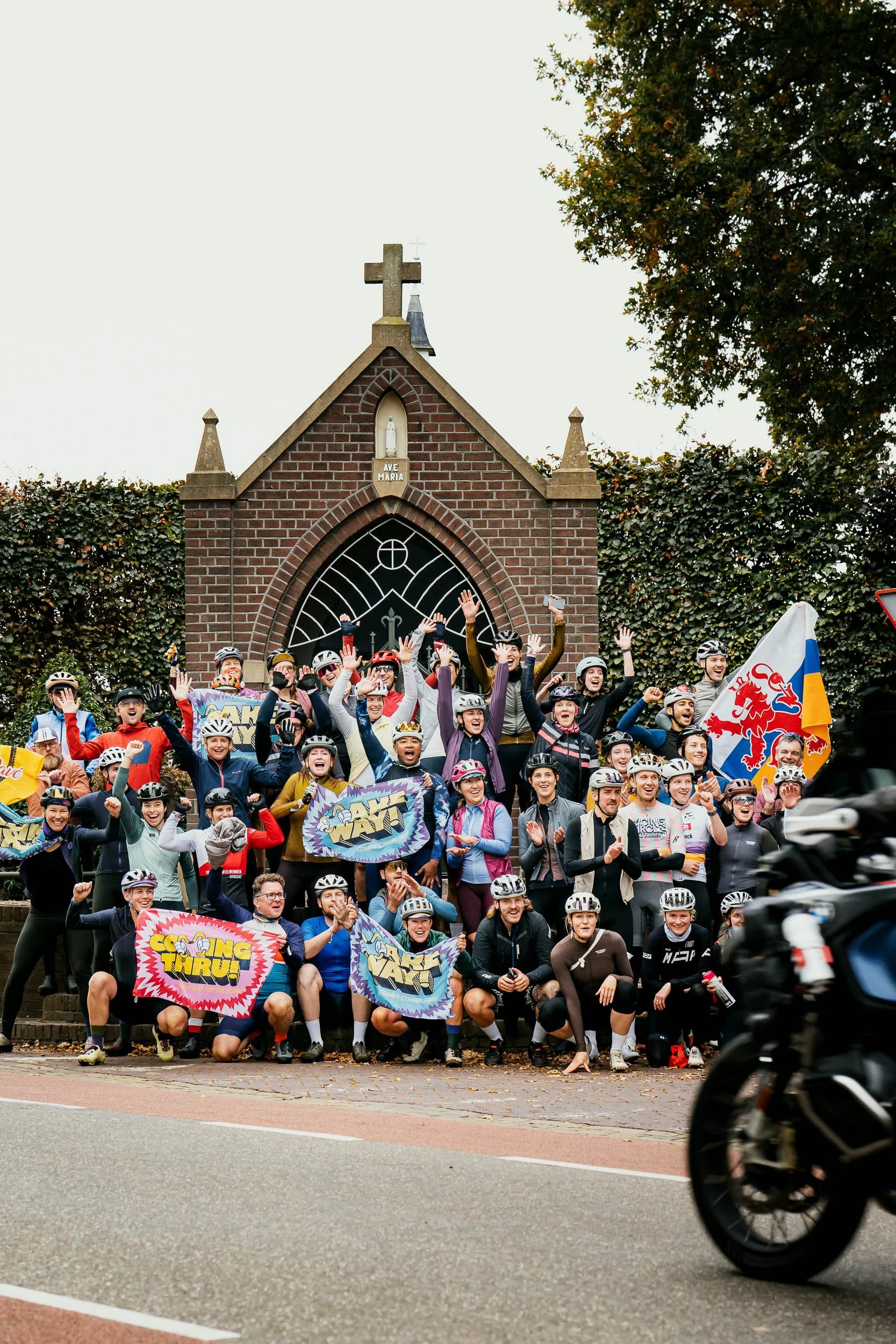 A large group of people with bicycle helmets, holding flags and signs, gathered in front of a small brick church with a cross on top, celebrating during daytime.