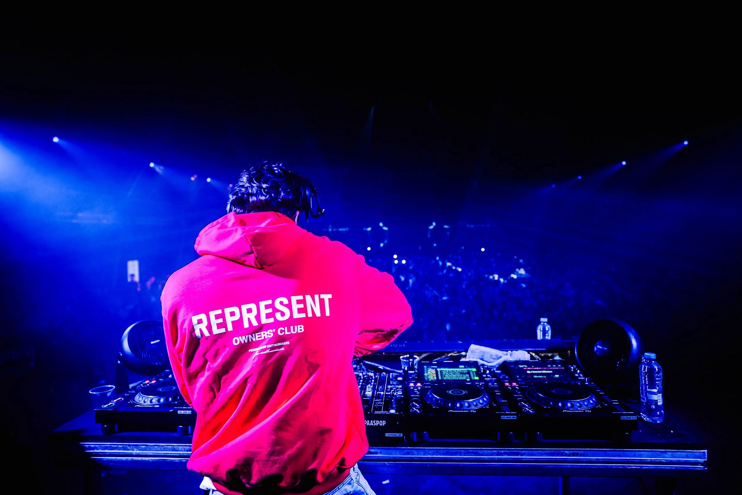 DJ performing on stage at a concert with dark background and blue lighting, wearing a red hoodie that says 'REPRESET OWNERS' CLUB' on the back. (Viktor Verhulst)
