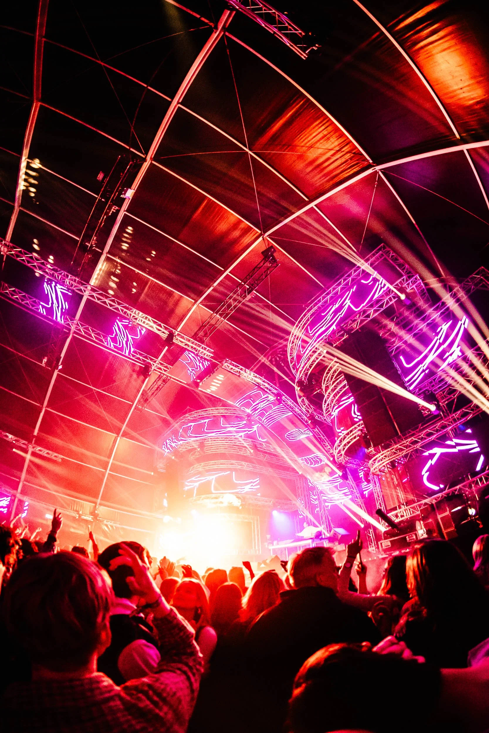 A lively concert scene inside a large venue with an elaborate lighting setup featuring purple and pink neon lights, with a crowd of people dancing and enjoying the show.  (Viktor Verhulst)