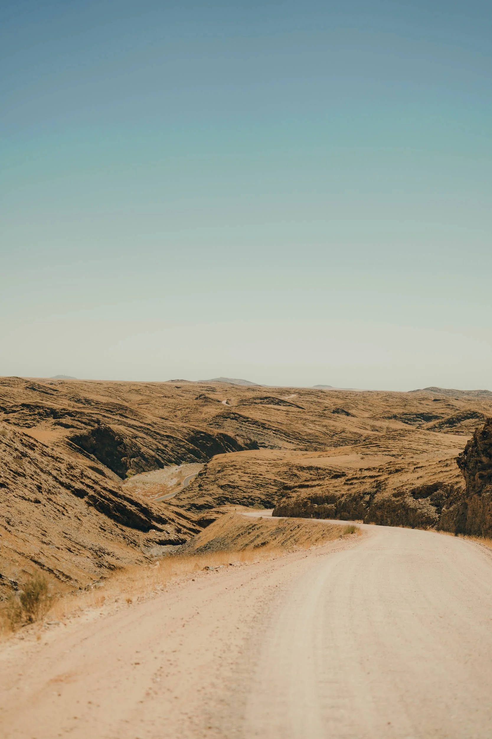 A winding dirt road running through dry, hilly terrain with sparse vegetation under a clear, blue sky.