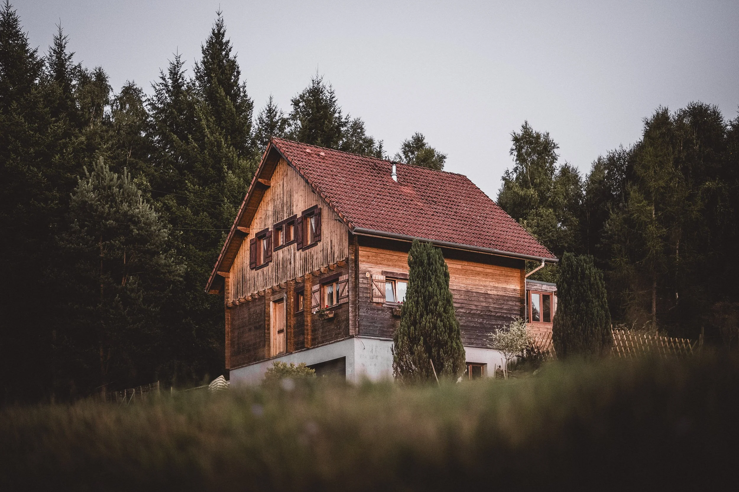 A wooden house with a red-tiled roof in a forested area, surrounded by trees and greenery.