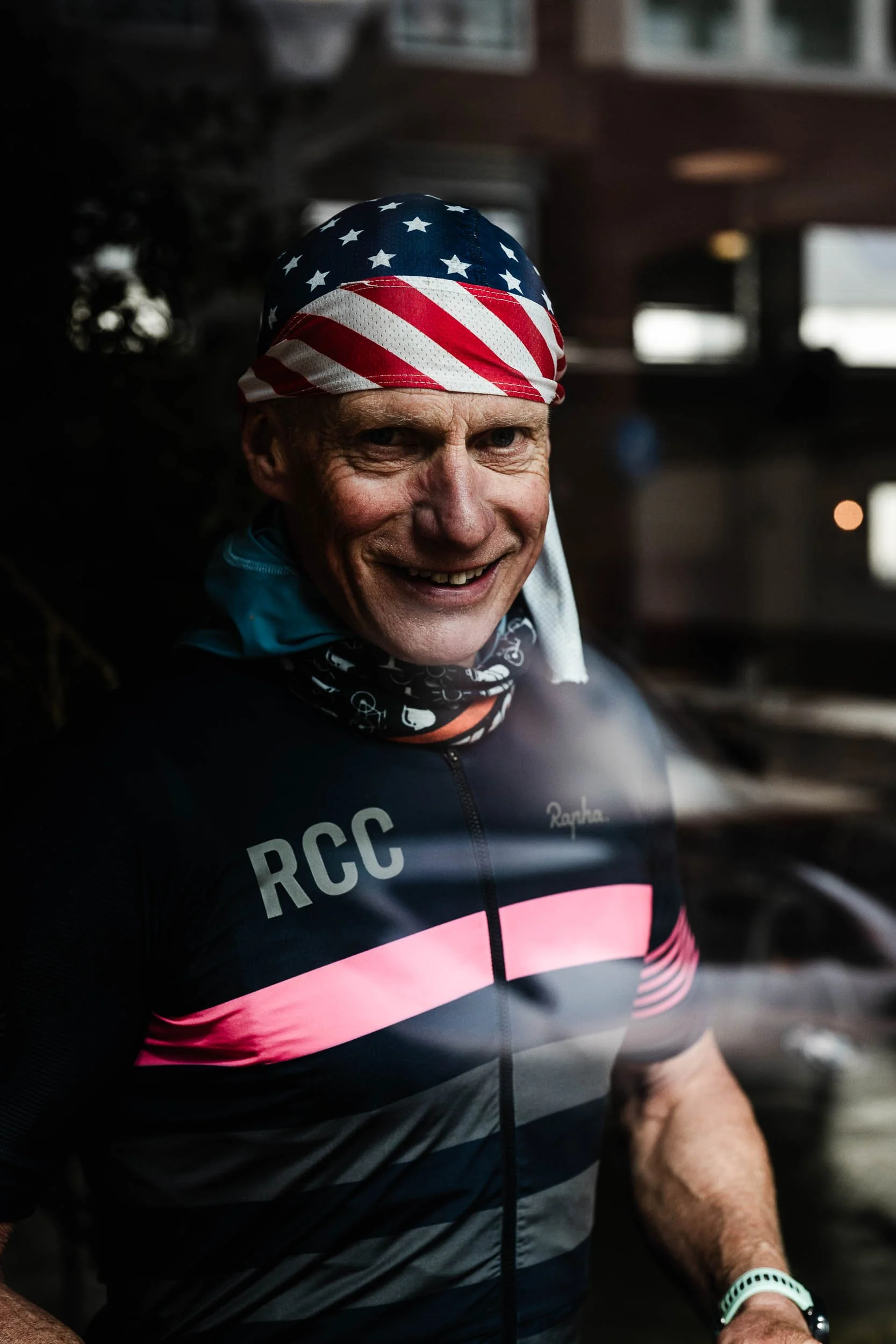 A man wearing a cycling jersey with 'RCC' and a pink stripe, smiling, with an American flag bandana on his head, in an indoor setting.