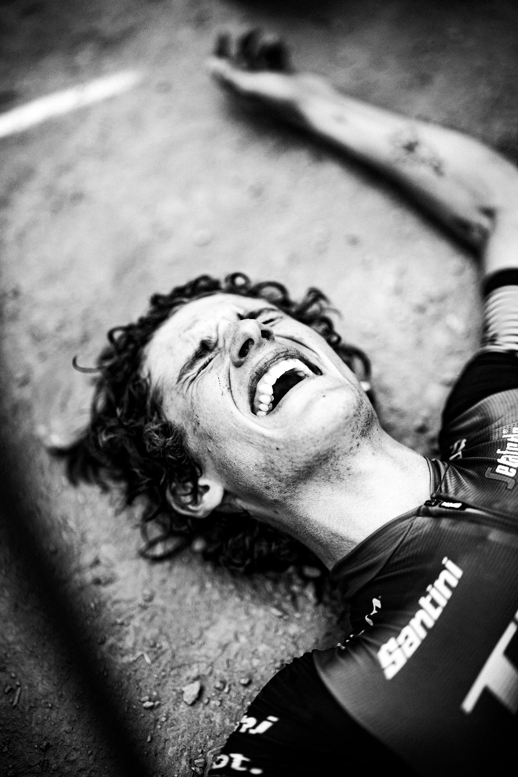 A cyclist is lying on the ground, visibly distressed or exhausted, with their eyes closed and mouth open, on a dirt or gravel surface.