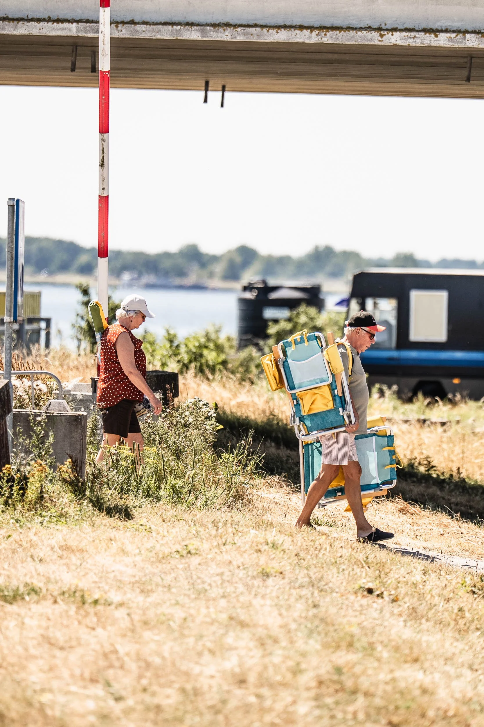 Two elderly people, a man and a woman, carrying yellow and blue lounge chairs and an umbrella, walking on a grassy path near a beach or lakeside with boats in the background. (Patta.nl Rapha)