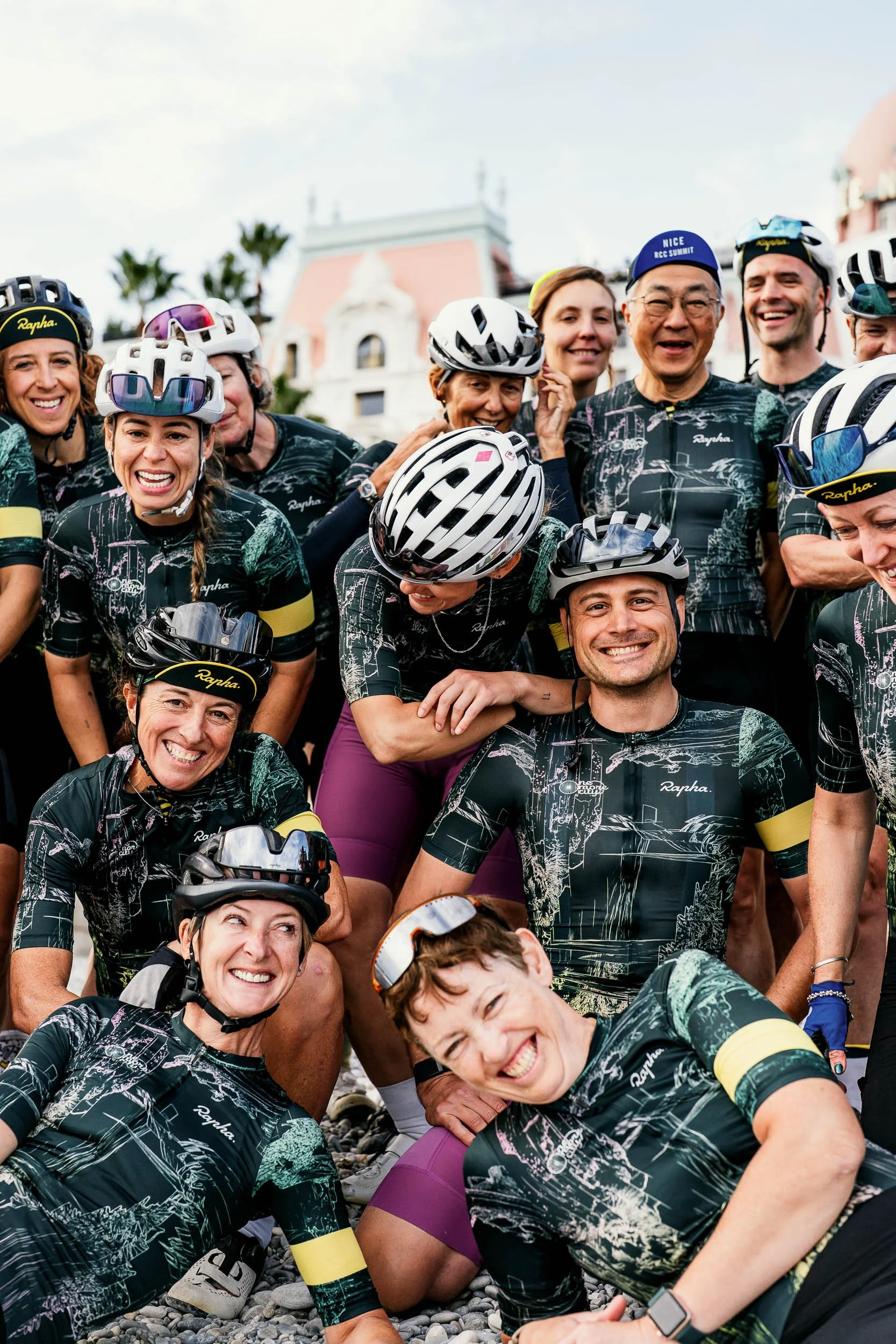 Group of smiling cyclists wearing matching jerseys and helmets, gathered outdoors in front of a historic building with palm trees. (One More City  Rapha.cc)