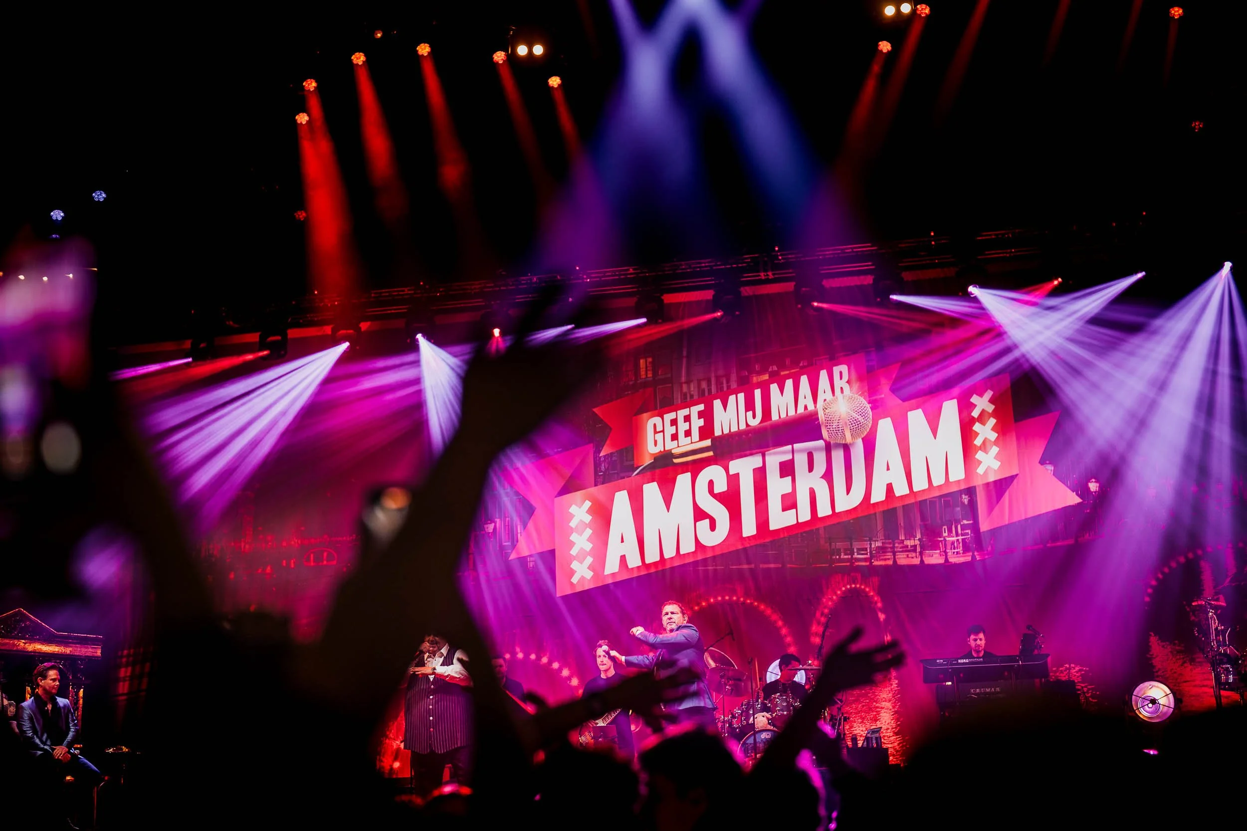 Concert stage with bright purple, red, and pink lights, a large screen displaying the words 'GEEF MIJ MAAR AMSTERDAM', a performer singing, and audience members raising their hands.