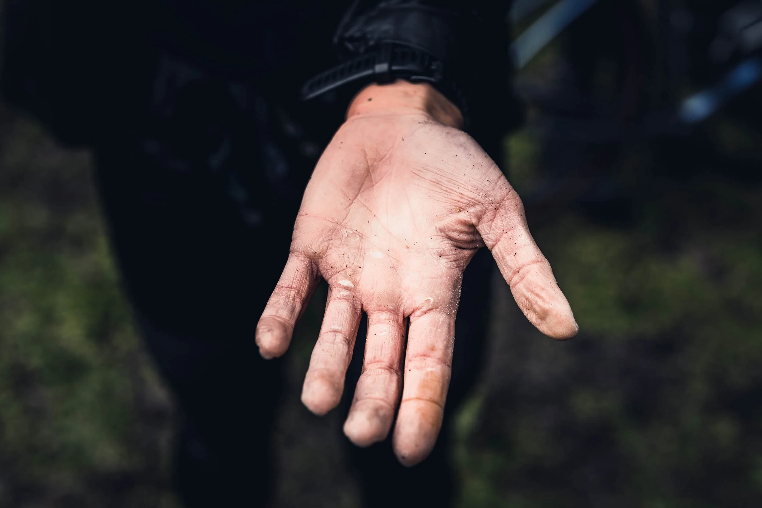 Close-up of a dirty and dusty human hand with a black wristwatch, outstretched with fingers slightly bent, against blurred outdoor background.