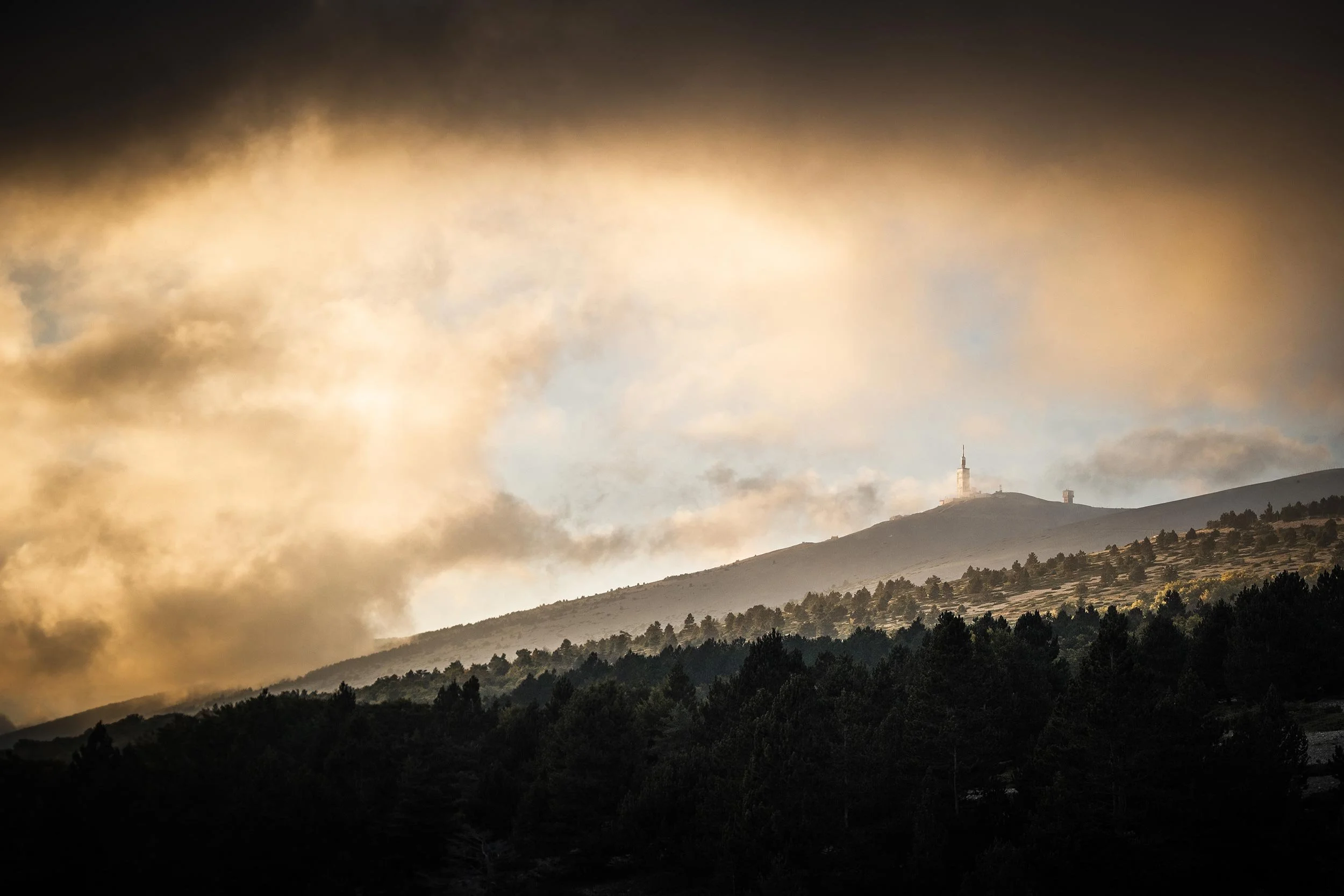 Mountain with trees in the foreground and a communication tower on the peak, with clouds in the sky.