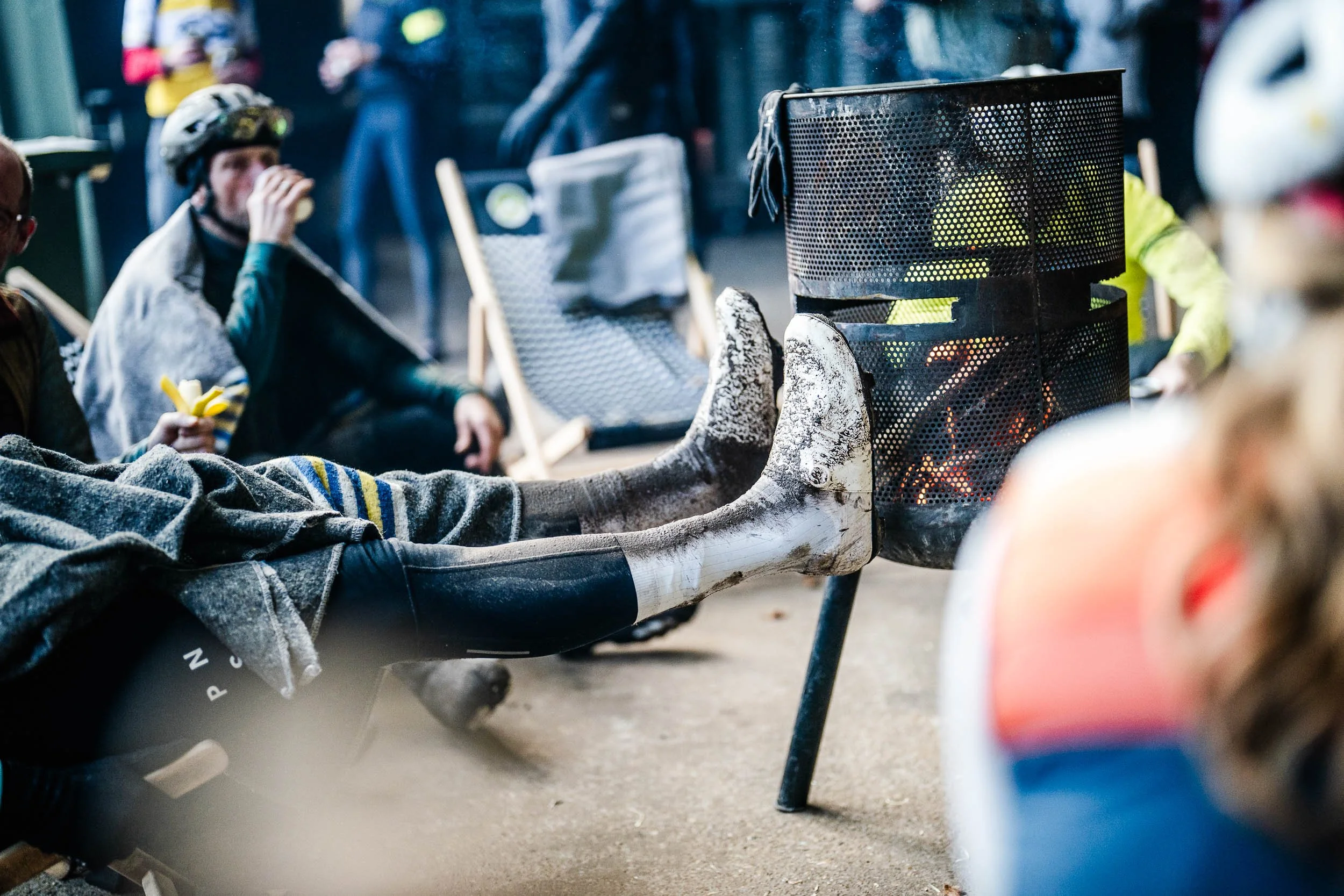 People sitting outdoors around a fire pit, with some wearing ski or snow boots and helmets, and others eating snacks, suggesting a casual social gathering.