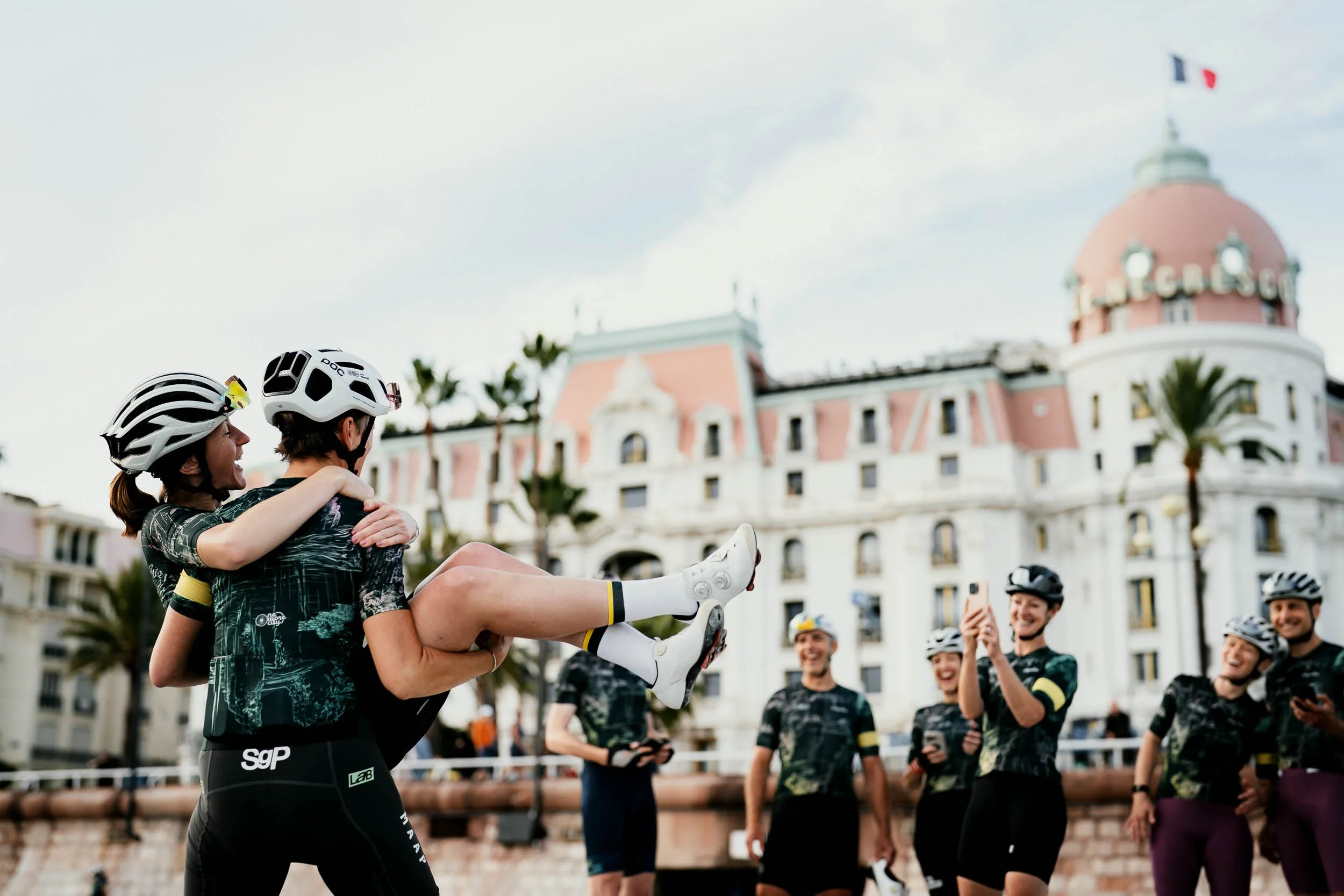 A group of cyclists practicing or celebrating in front of a historic building, with two women wearing helmets smiling and hugging, while others in the background take pictures and laugh. (One More City  Rapha.cc)