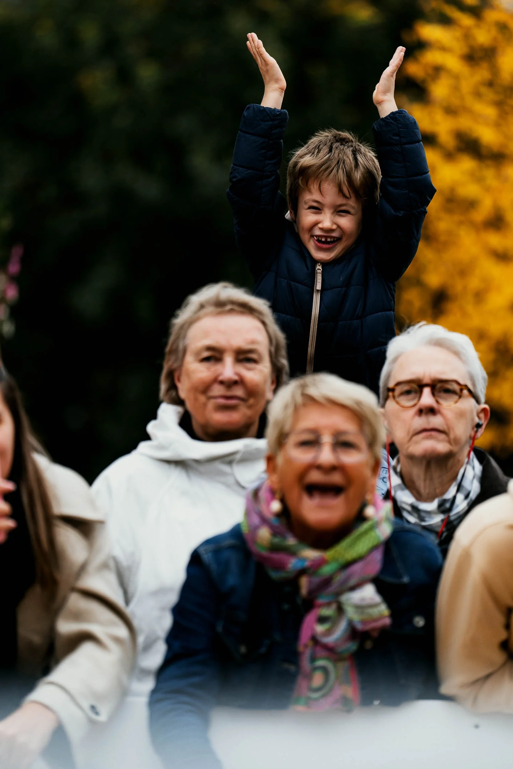 Young boy with brown hair in a dark blue jacket smiling and raising his hands in the air while sitting on an adult's shoulders, with a group of older adults and a woman with glasses and a colorful scarf in outdoor setting with fall foliage.
