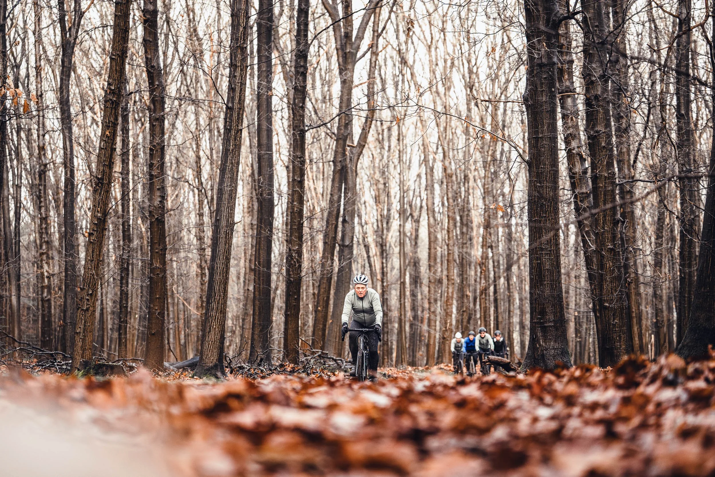 A person riding a bicycle through a leaf-covered forest with several other cyclists in the background on a cloudy day.