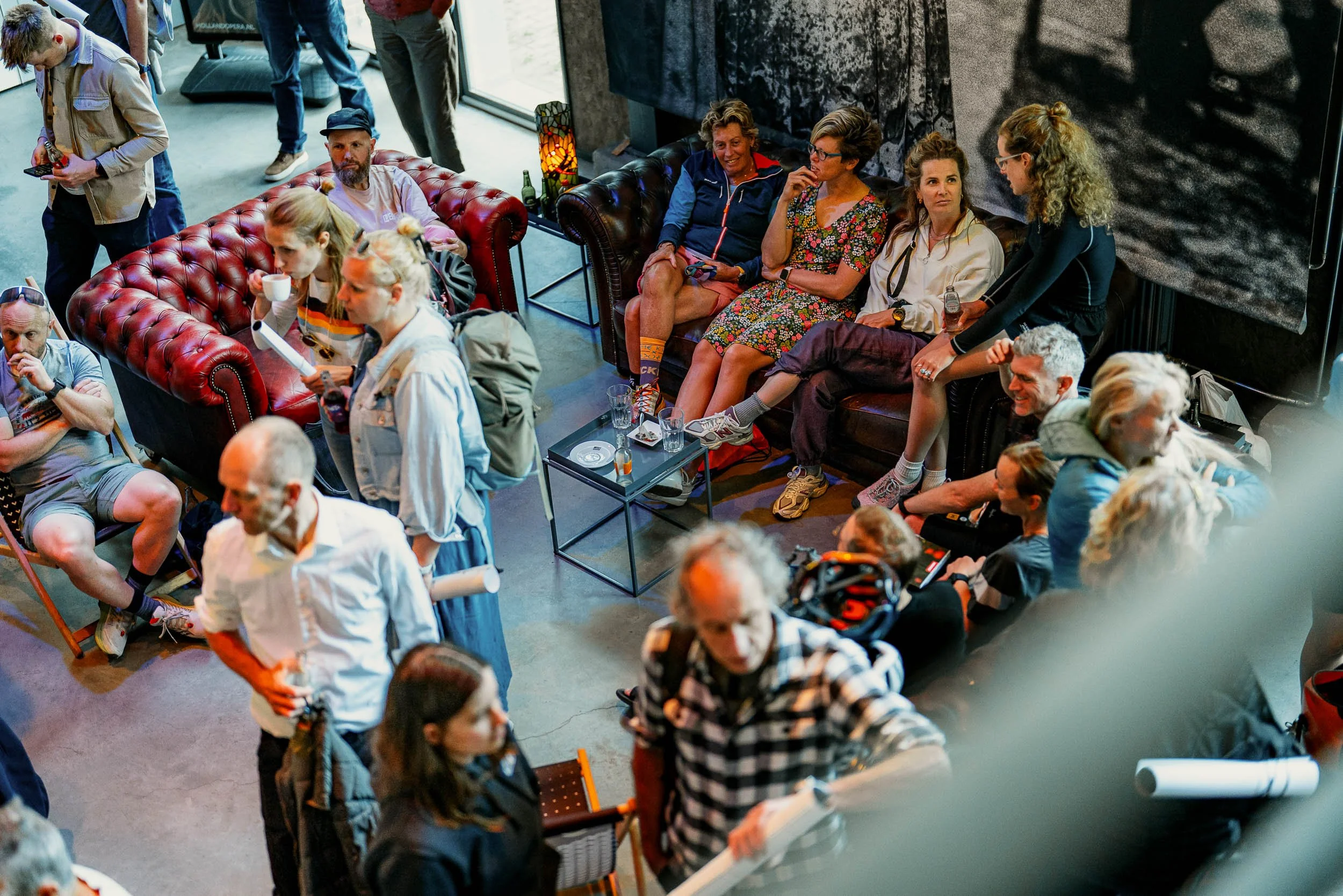 People gathered in a modern indoor space with leather couches, some sitting and some standing, engaging in conversations and looking at their phones.