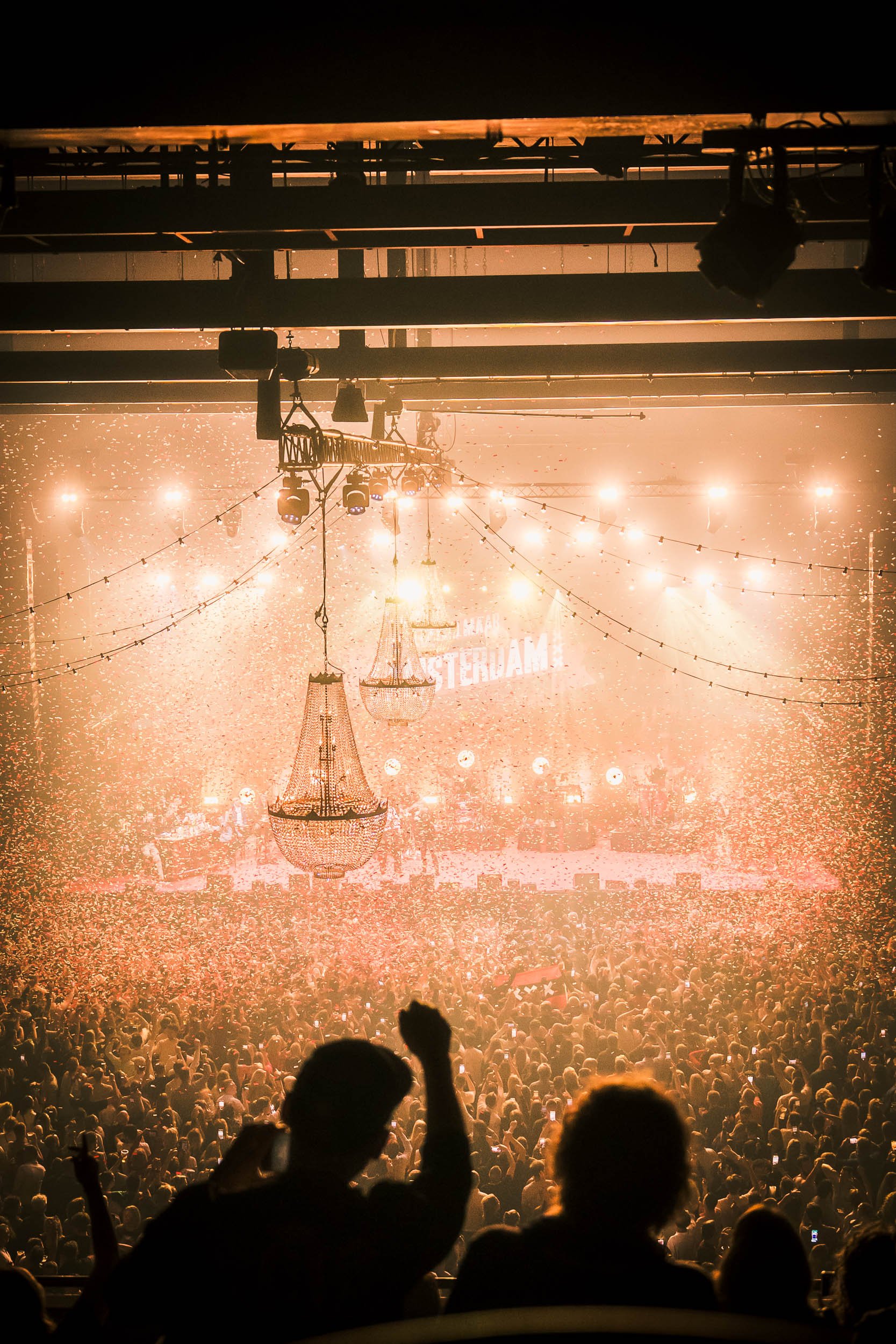 Concert venue with large crowd, stage, chandeliers, and bright lights, celebrating in Amsterdam.  (Amsterdam / Ziggo)