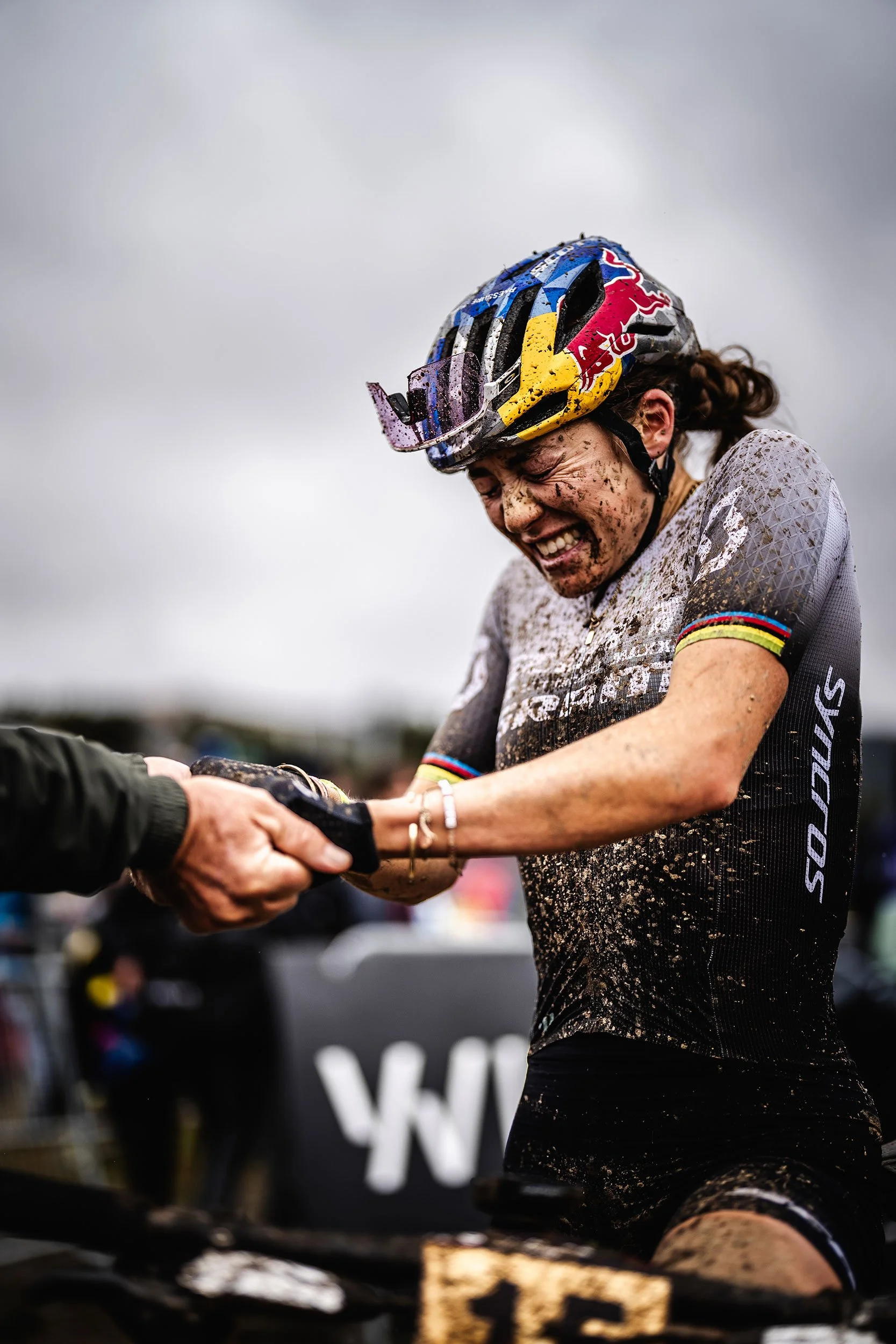 A female cyclist with a muddy face and jersey shaking hands with someone after a race, wearing a helmet and smiling.