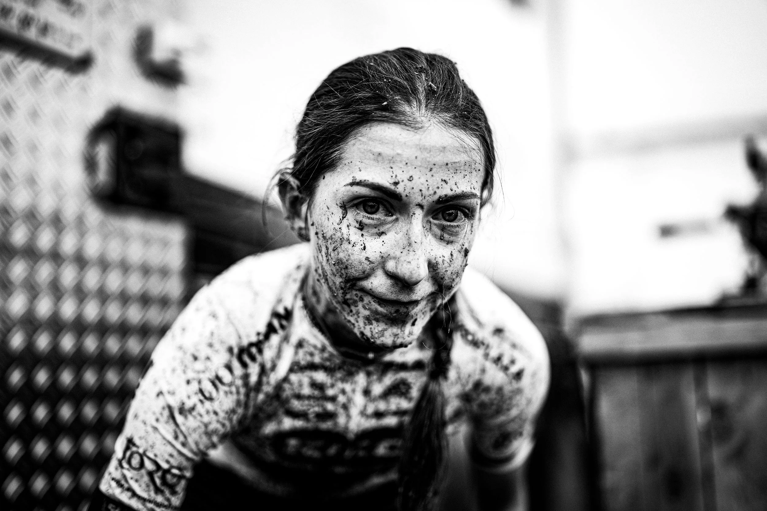 A woman with dark hair in a braid, covered in mud or paint, leaning forward and looking at the camera in an indoor setting.