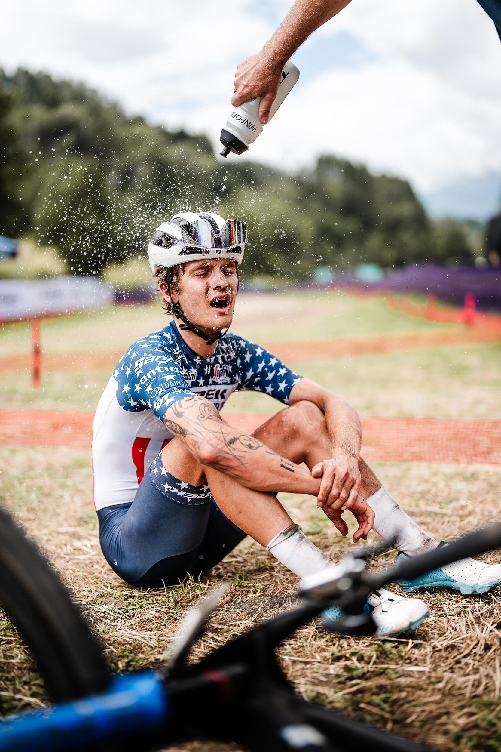 A cyclist sitting on the ground in distress after a crash during a race, receiving water from a person off-camera.