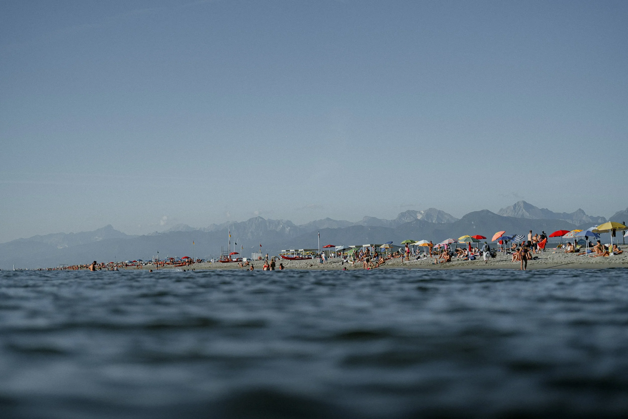 Beach scene with people under colorful umbrellas, sandy shore with some boats, and mountains in the background on a clear day.