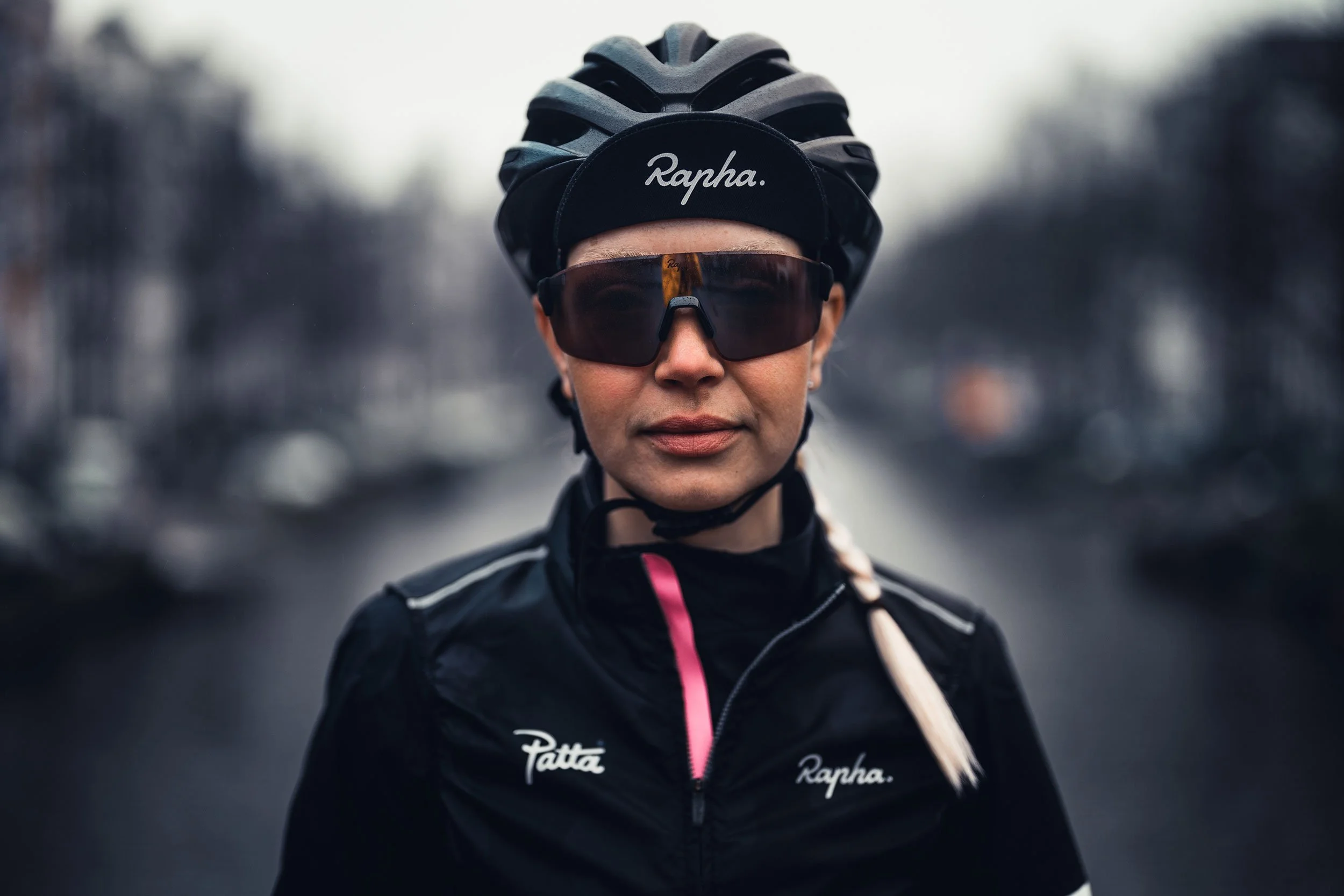 A woman wearing a bike helmet, large sunglasses, and a black sports jacket standing outdoors on a cloudy day.