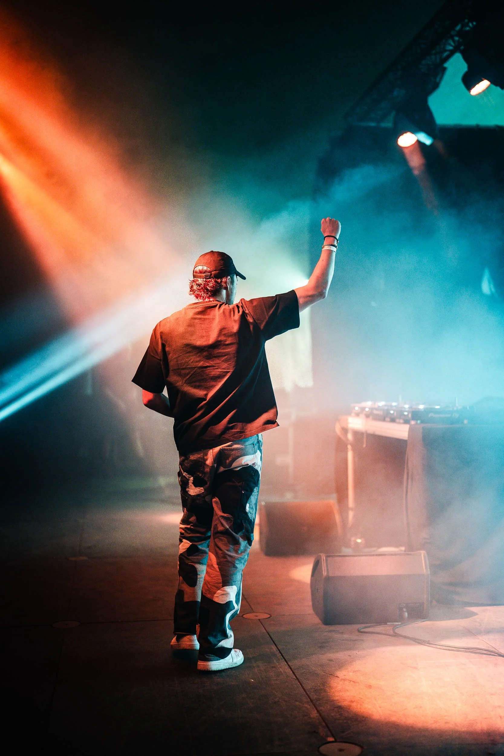 A person with curly hair, wearing a cap, a black t-shirt, and camo pants, performing on stage with one arm raised, surrounded by colorful stage lighting and fog.   (Viktor Verhulst)