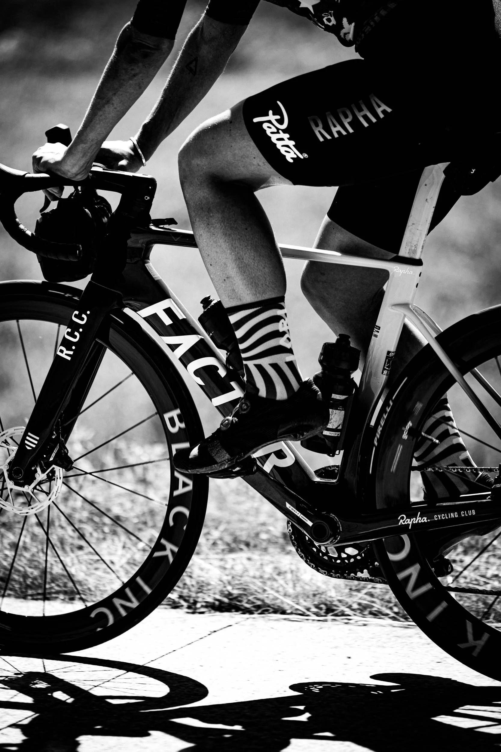 A cyclist riding a racing bike on a paved path, with onlookers in the background. The image is in black and white. (Patta.nl Rapha)