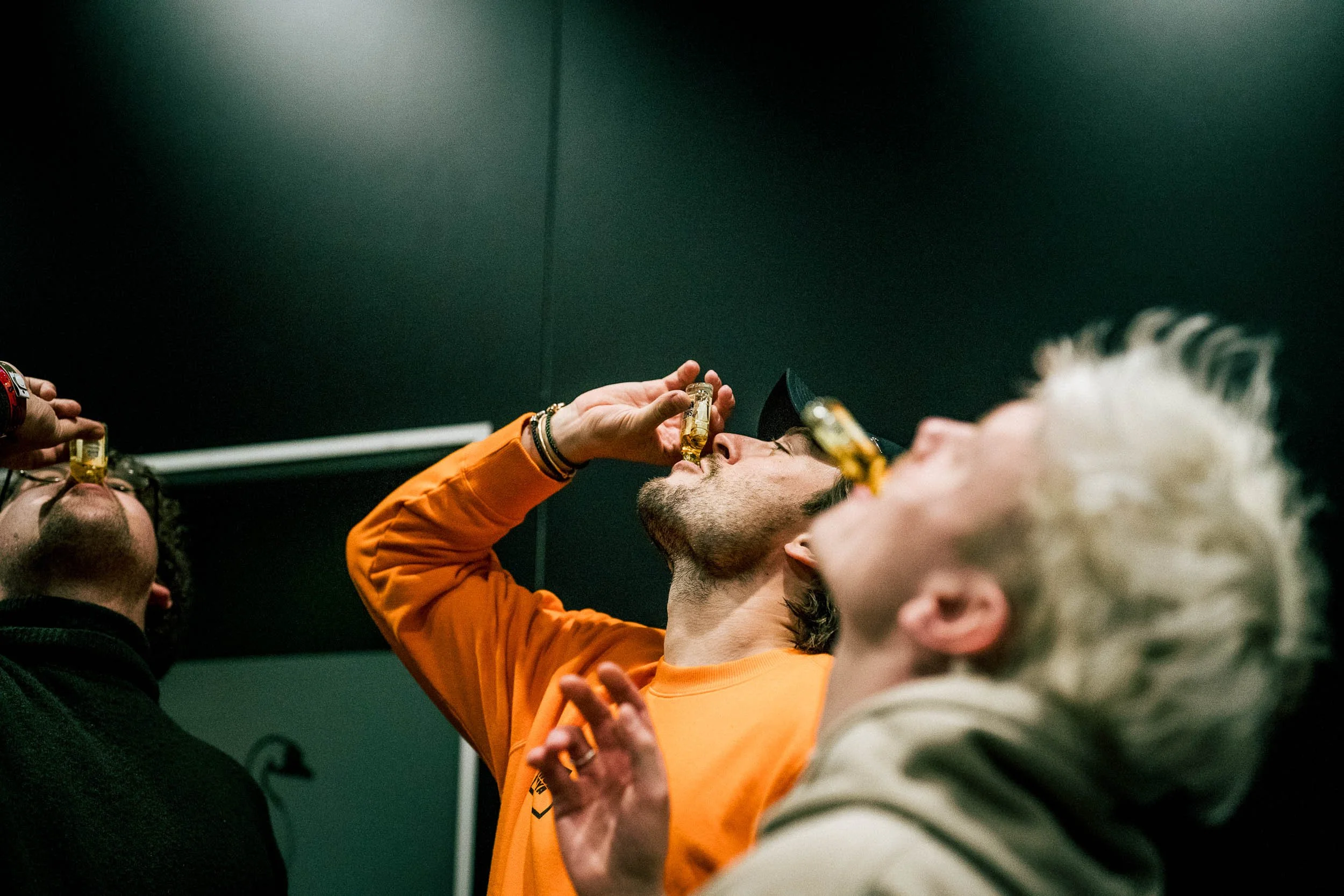 Three people are taking shots of alcohol, with two of them tilting their heads back and one holding a bottle to his nose.