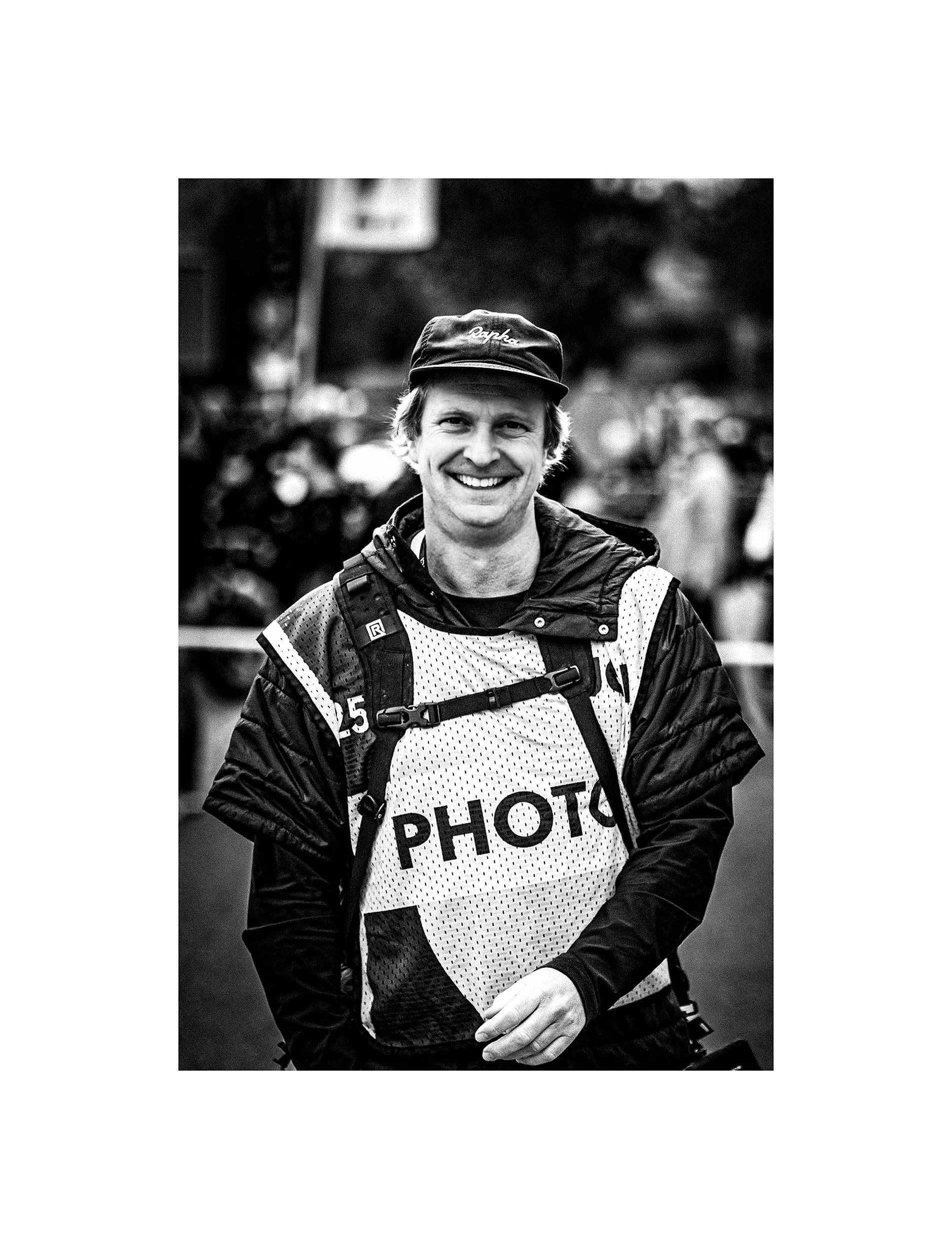 Black and white photo of a smiling man wearing a cap, a jacket, and a vest labeled 'PHOTO,' with a backpack, at an outdoor event. (Vincent Engel Fotografie)