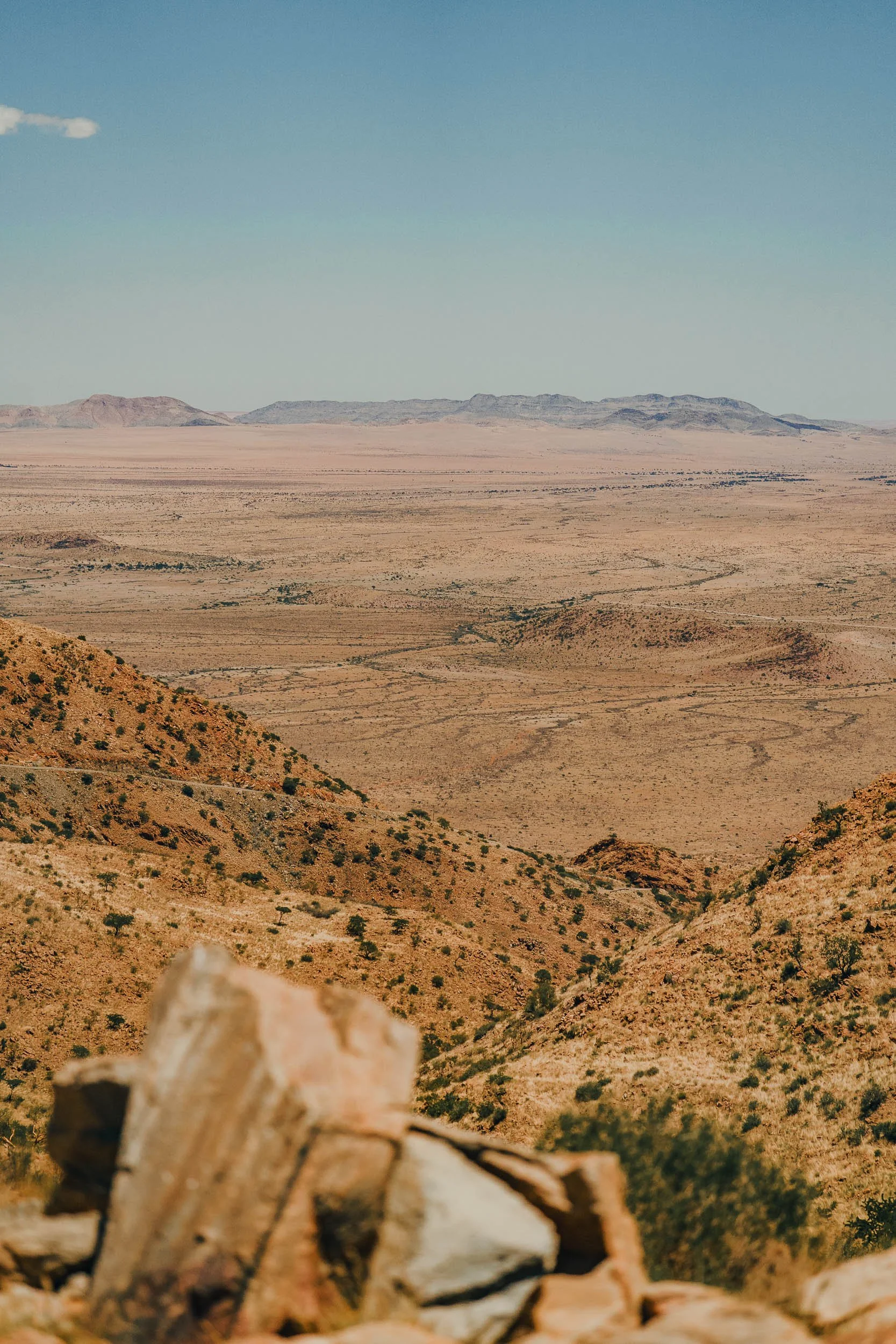 Arid desert landscape with rocky hills in the foreground and flat plains with mountain ranges in the background under a clear blue sky.