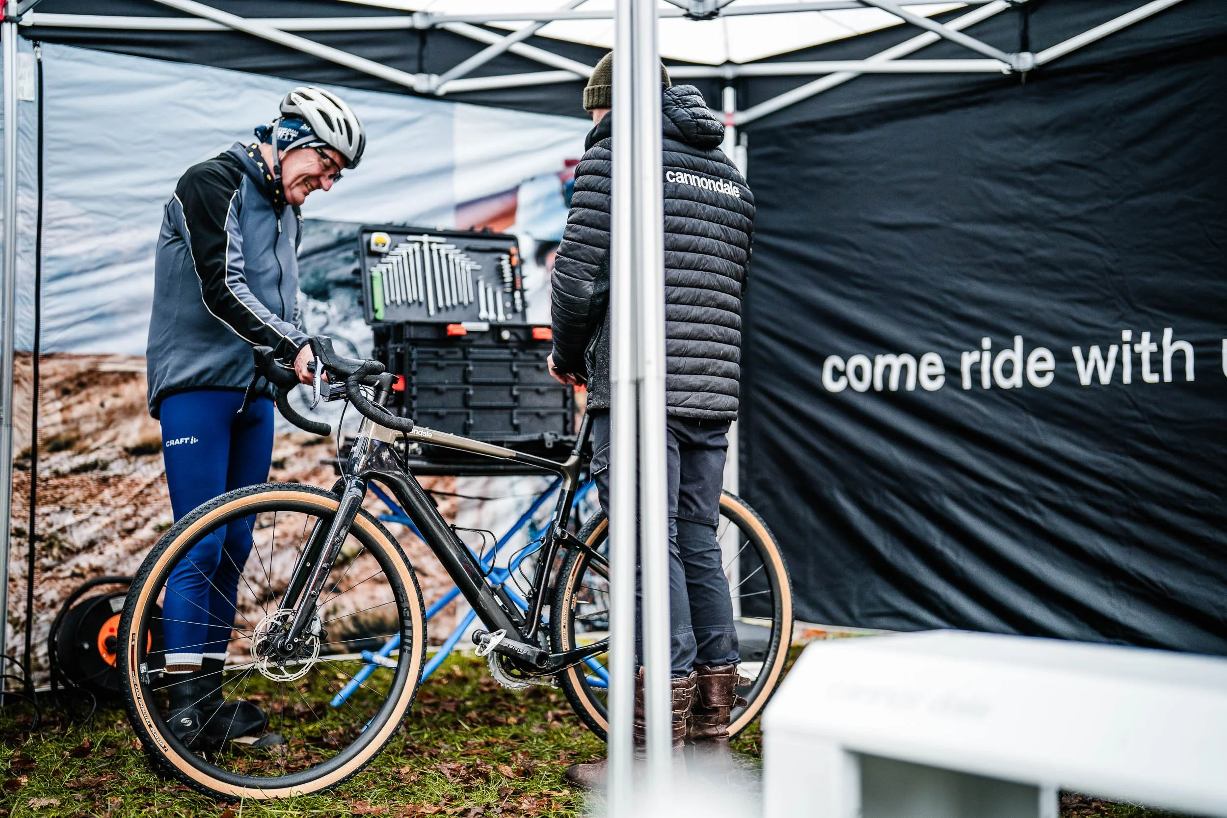 Two men working on a black bicycle inside a black canopy tent, with one man wearing a helmet and the other wearing a black jacket with 'Cannondale' on the back, and a sign that says 'come ride with'.