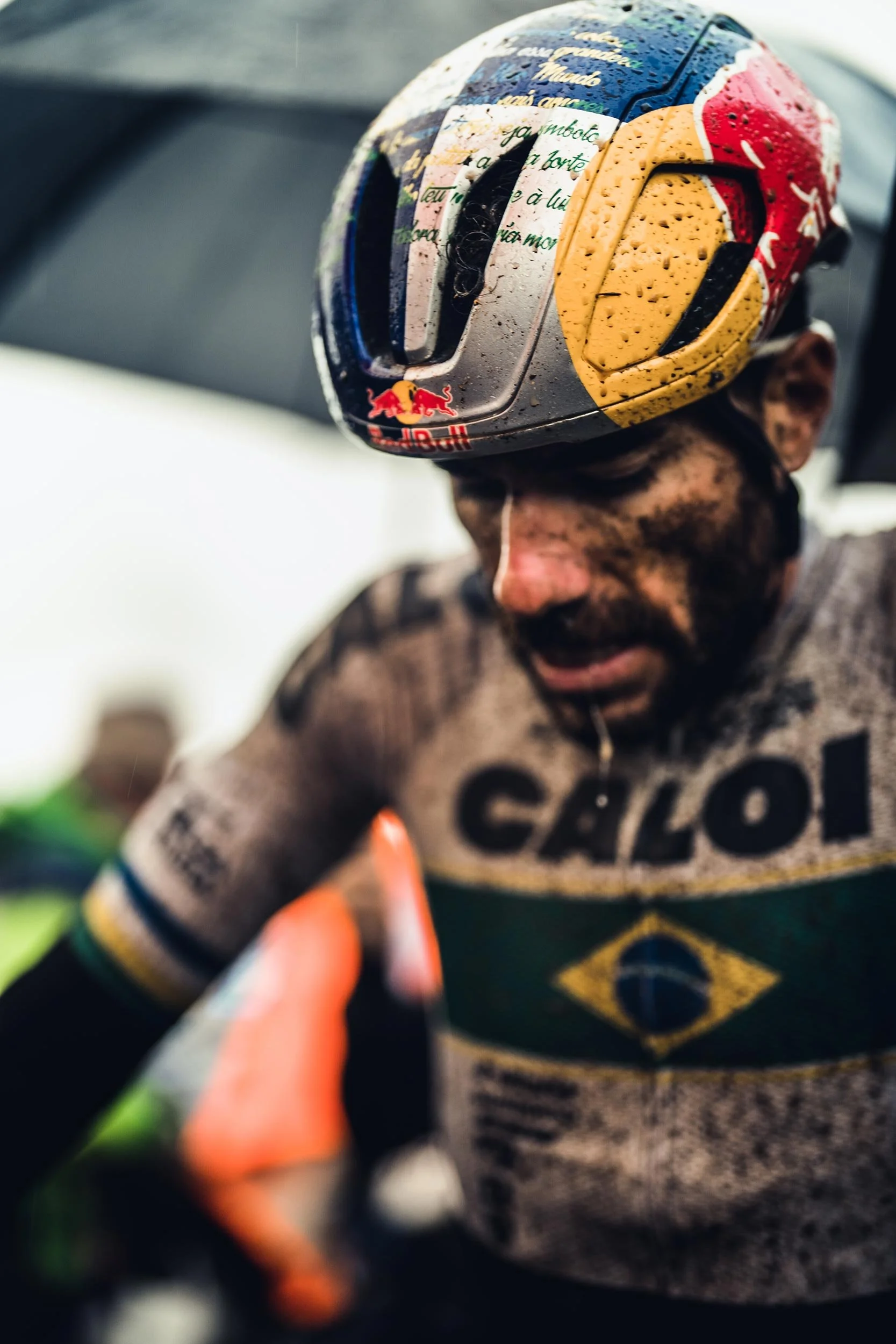 A cyclist with mud on his face and clothing wearing a helmet with the Red Bull logo during a race.