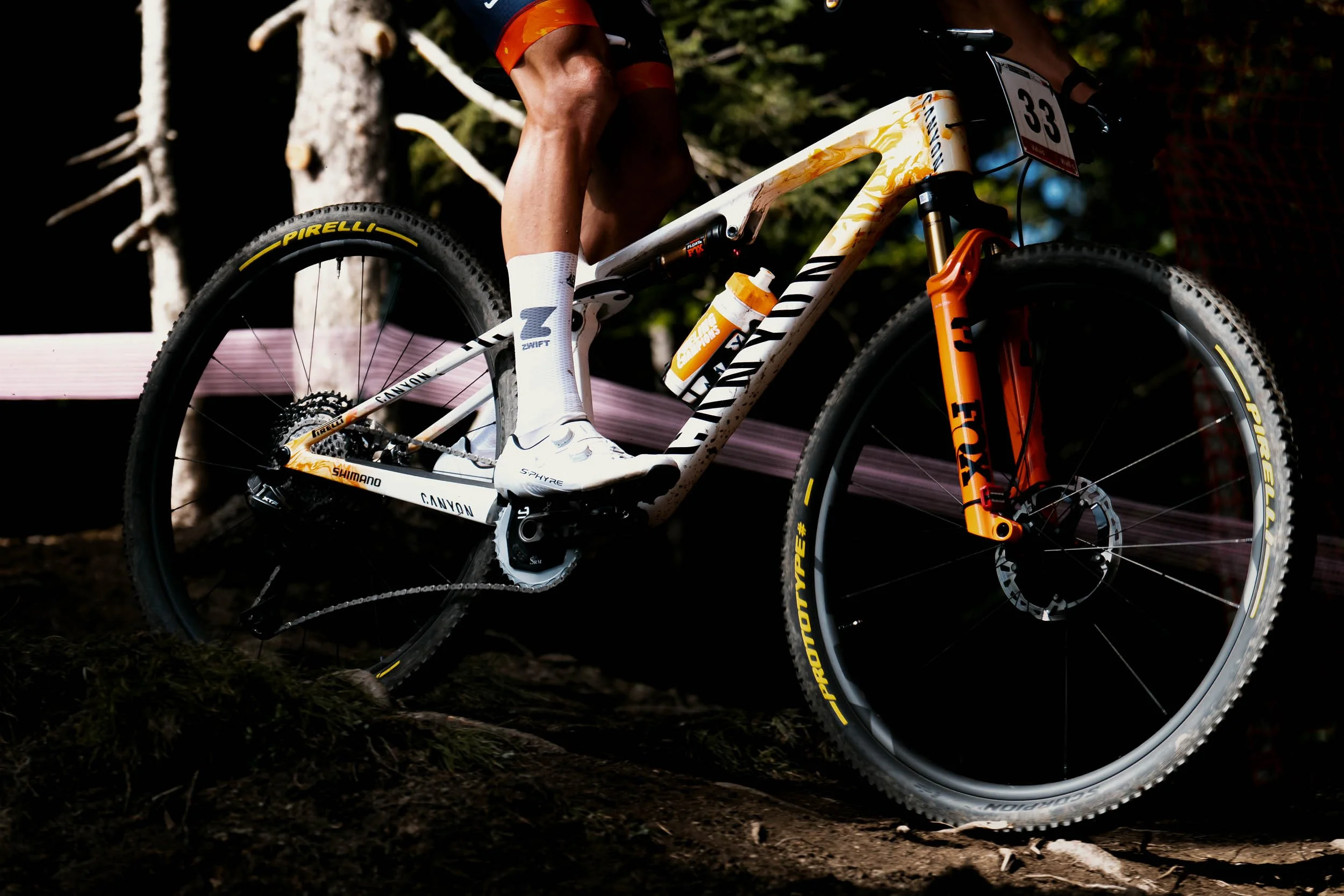 Close-up of a mountain biker riding on rough terrain, focusing on the bike's frame and wheels. The bike has a white and orange design, with Pirelli tires and FOX suspension. The rider wears white socks and shorts.