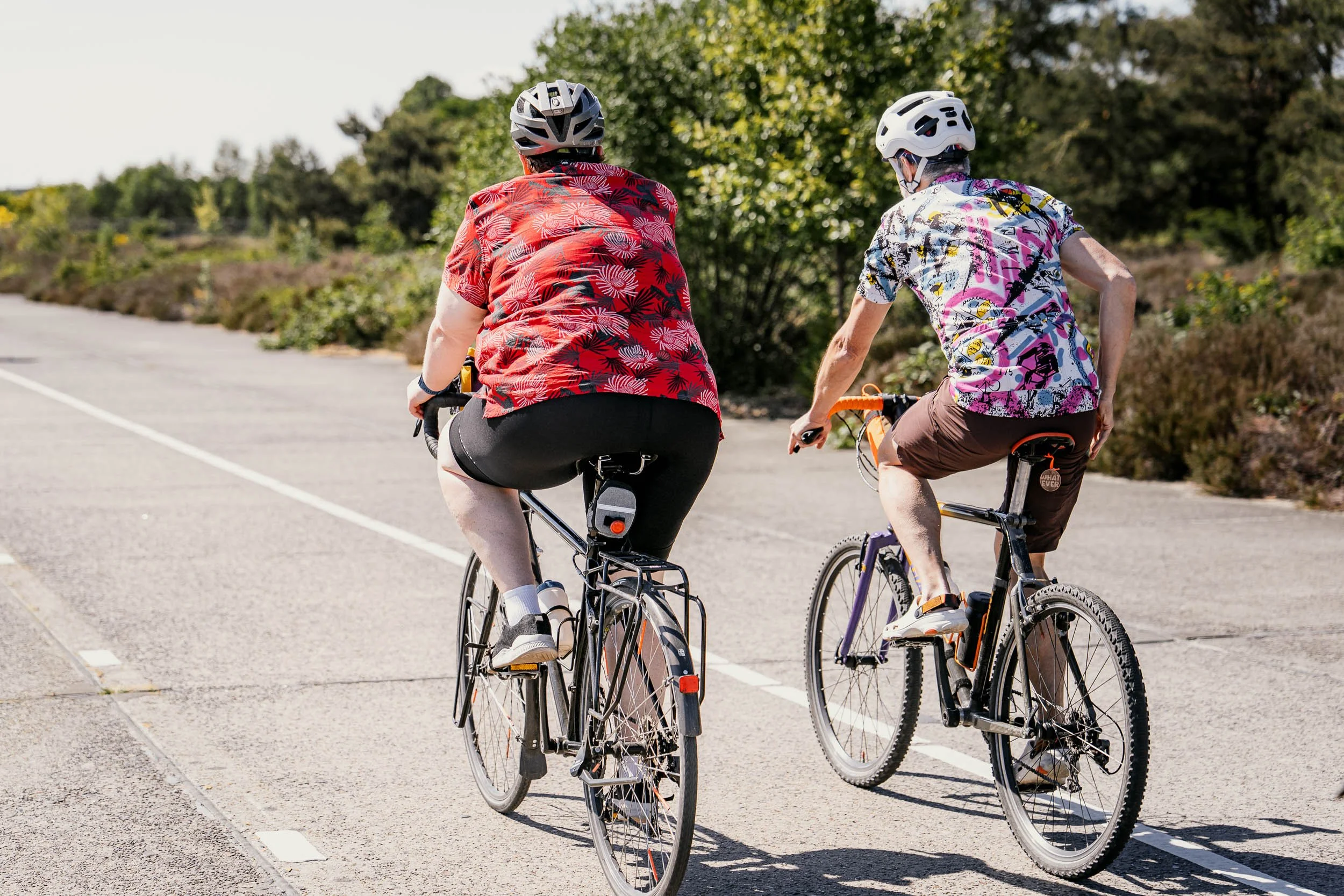 Two people riding bicycles on a paved path outdoors during daytime, wearing helmets and colorful shirts, with greenery and trees in the background.