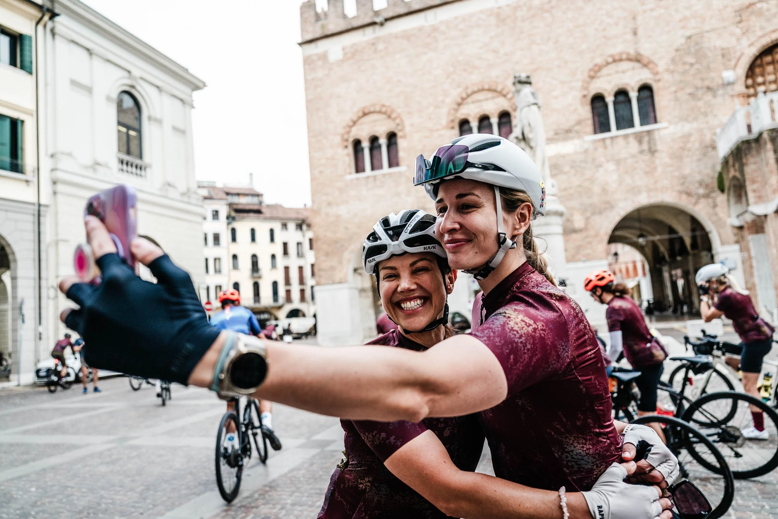 Two women in cycling gear, wearing helmets, smiling, and hugging while taking a selfie on a city street with other cyclists in the background and historic buildings.