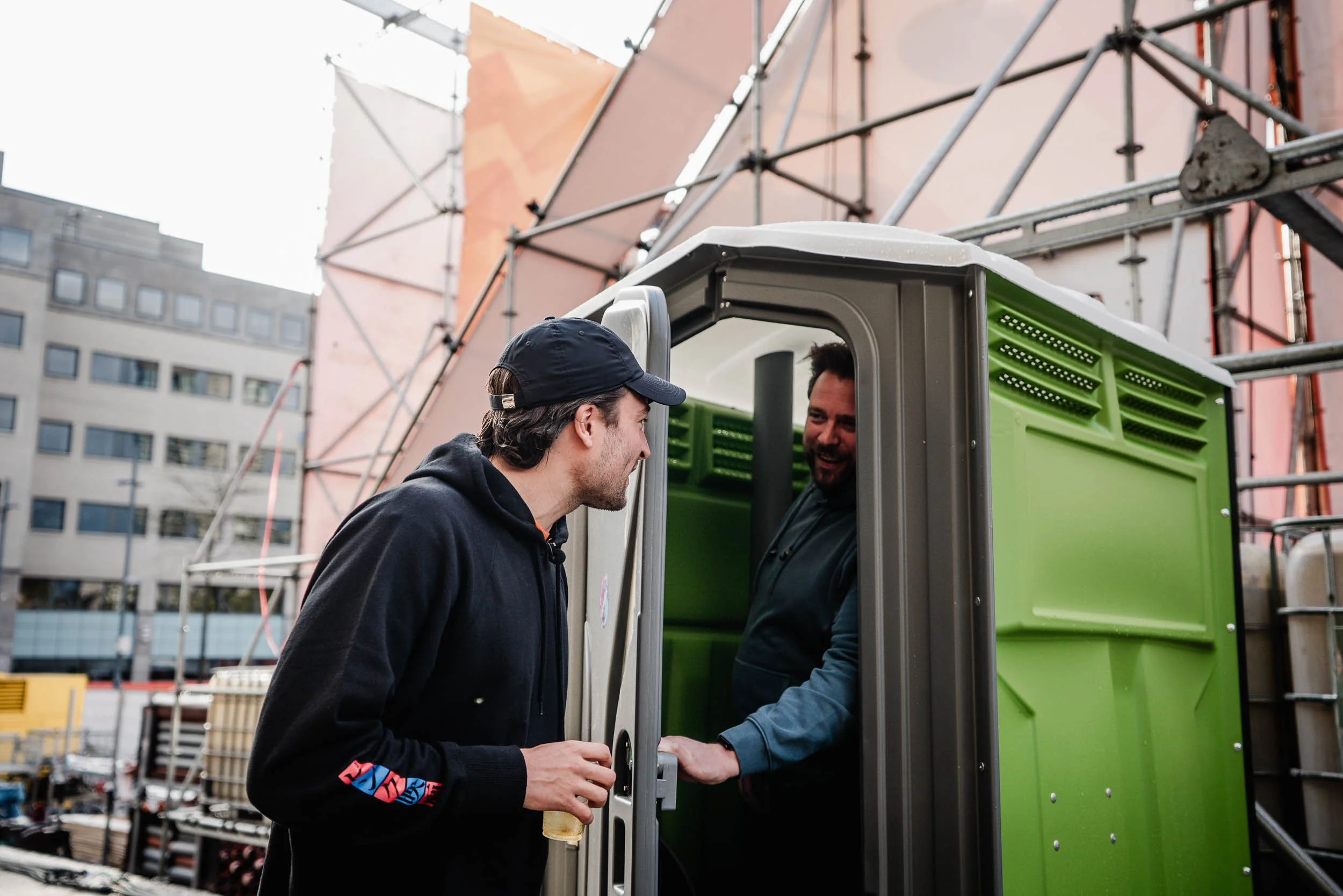 Two men talking at a construction site, one inside a green portable toilet and the other outside holding a beer.  (Viktor Verhulst)