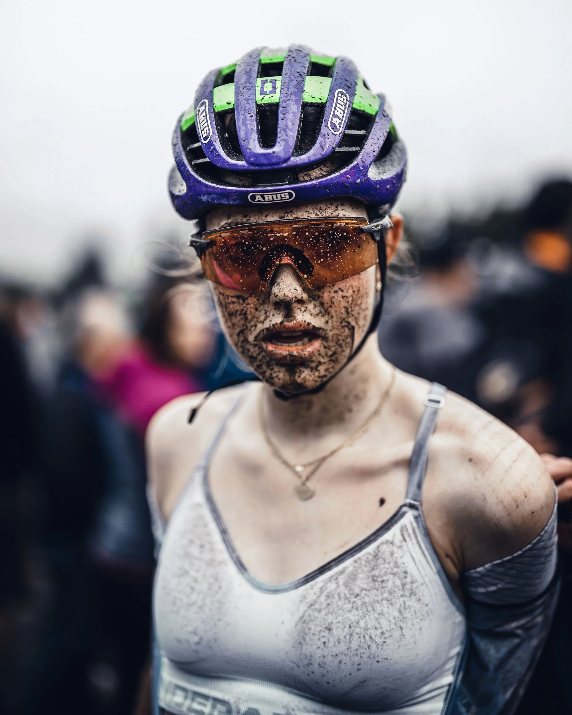 A female cyclist covered in mud, wearing a purple and green helmet and orange-tinted sunglasses.