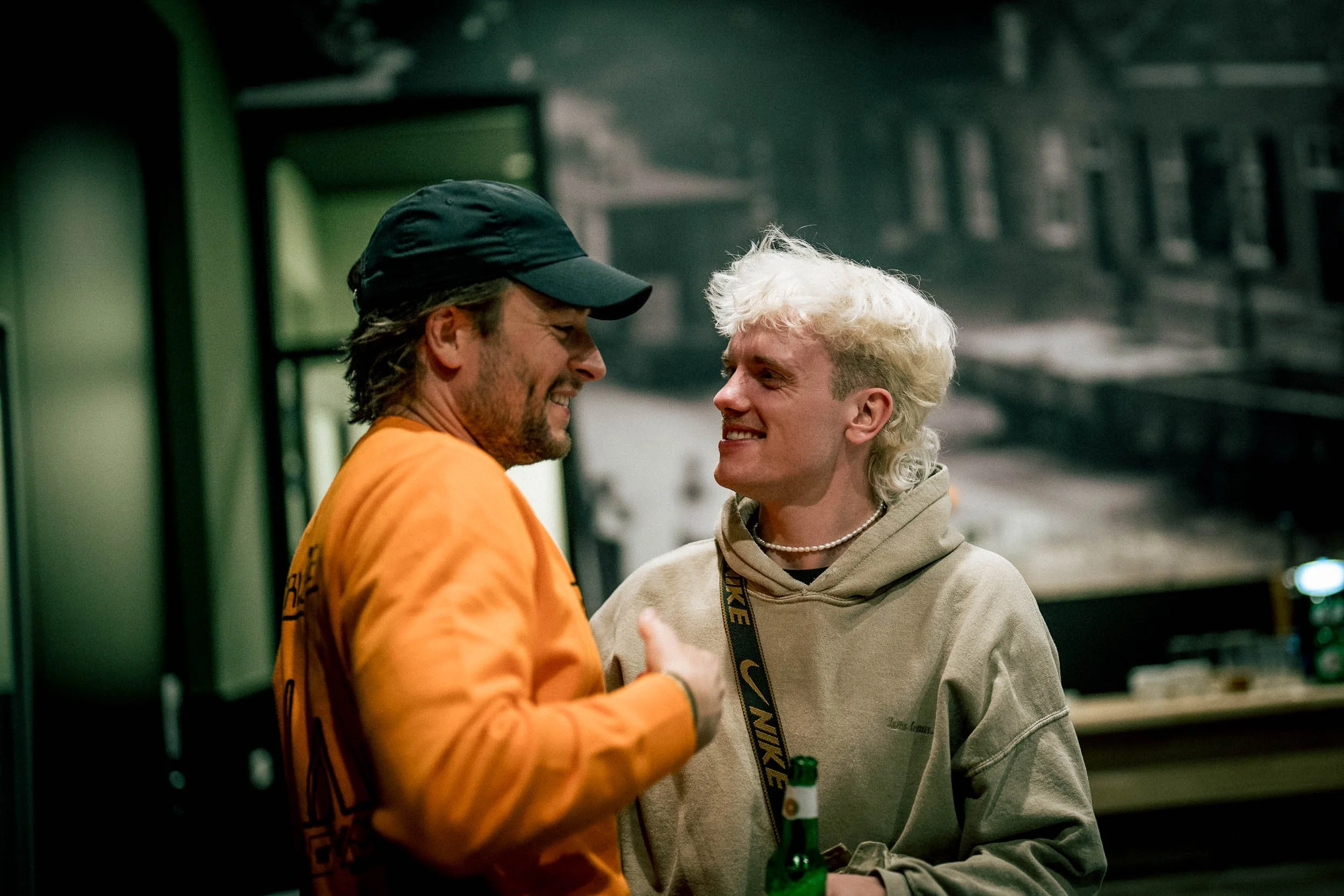 Two young men talking and smiling in a casual indoor setting, one with a cap and orange shirt, the other with curly blonde hair, beige hoodie, and a pearl necklace, holding a beer.   (Viktor Verhulst)