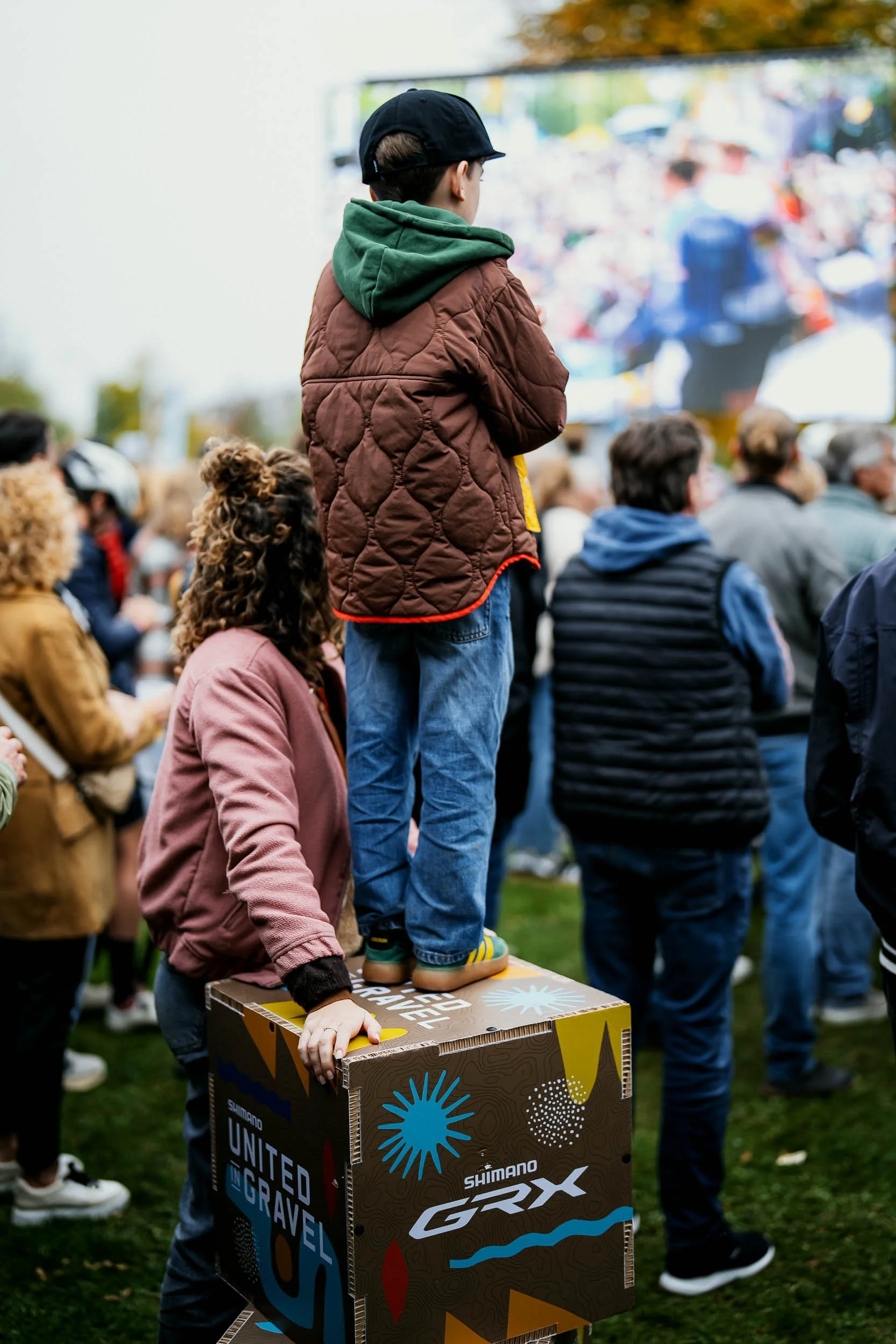 A child standing on a cardboard box at an outdoor event, surrounded by a group of people, with a large screen in the background displaying a crowd.