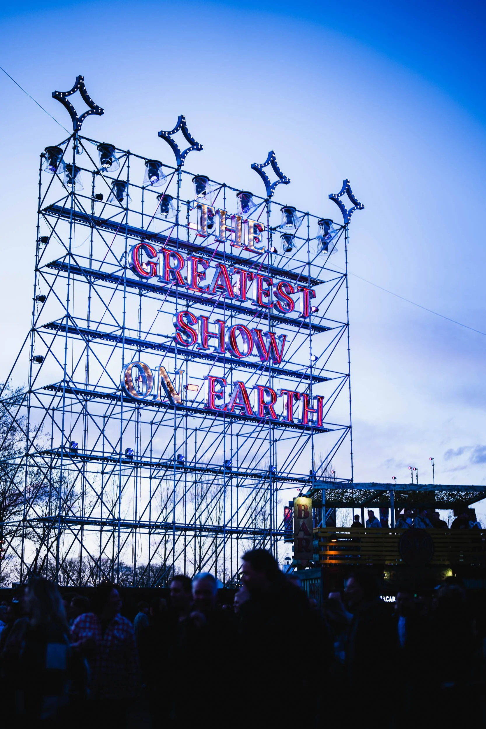 An outdoor carnival scene at dusk with a large illuminated sign reading 'The Greatest Show on Earth' on a tall metal structure, and people gathered below.  (Viktor Verhulst)