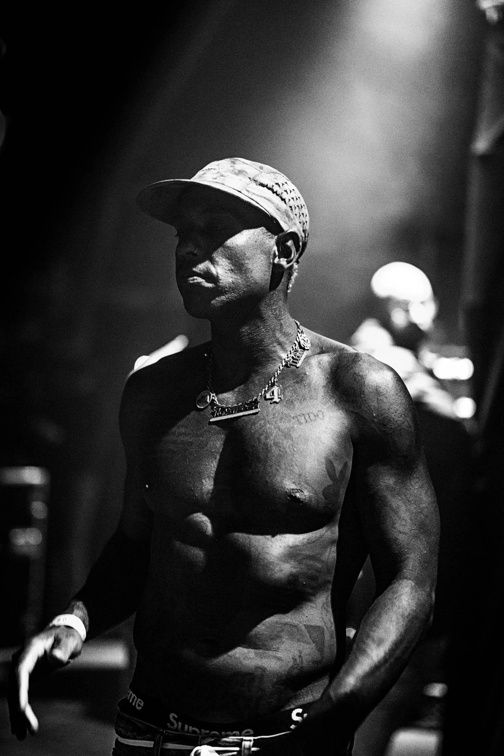 Black and white photo of shirtless man wearing a cap, chain necklace, and Supreme underwear, standing in a dimly lit environment. (Paaspop Bizzy)