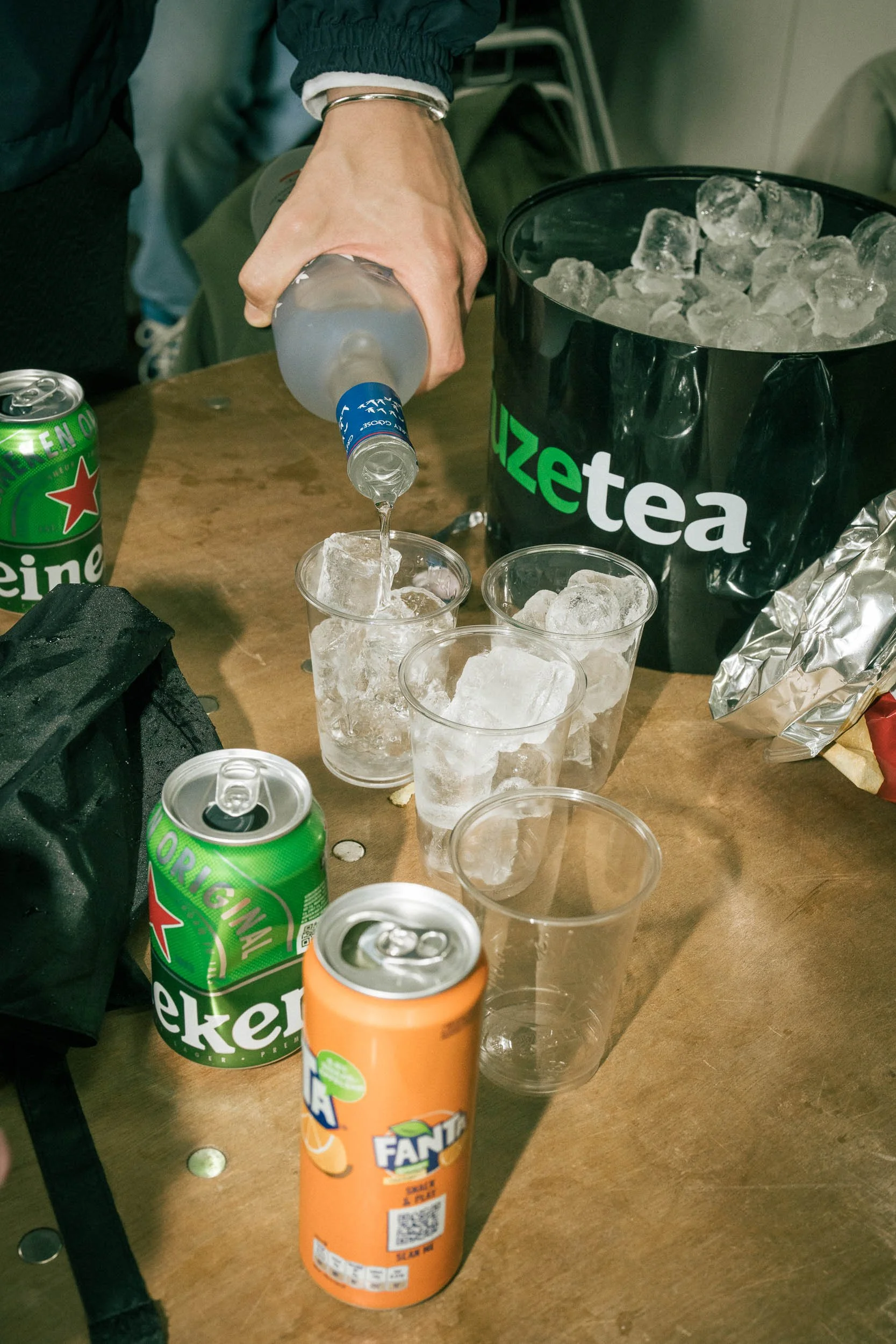 Person pouring water into a glass with ice on a wooden table, surrounded by cans of Heineken, Fanta, and a bucket of ice, with other empty glasses and a black bag nearby.  (Viktor Verhulst)