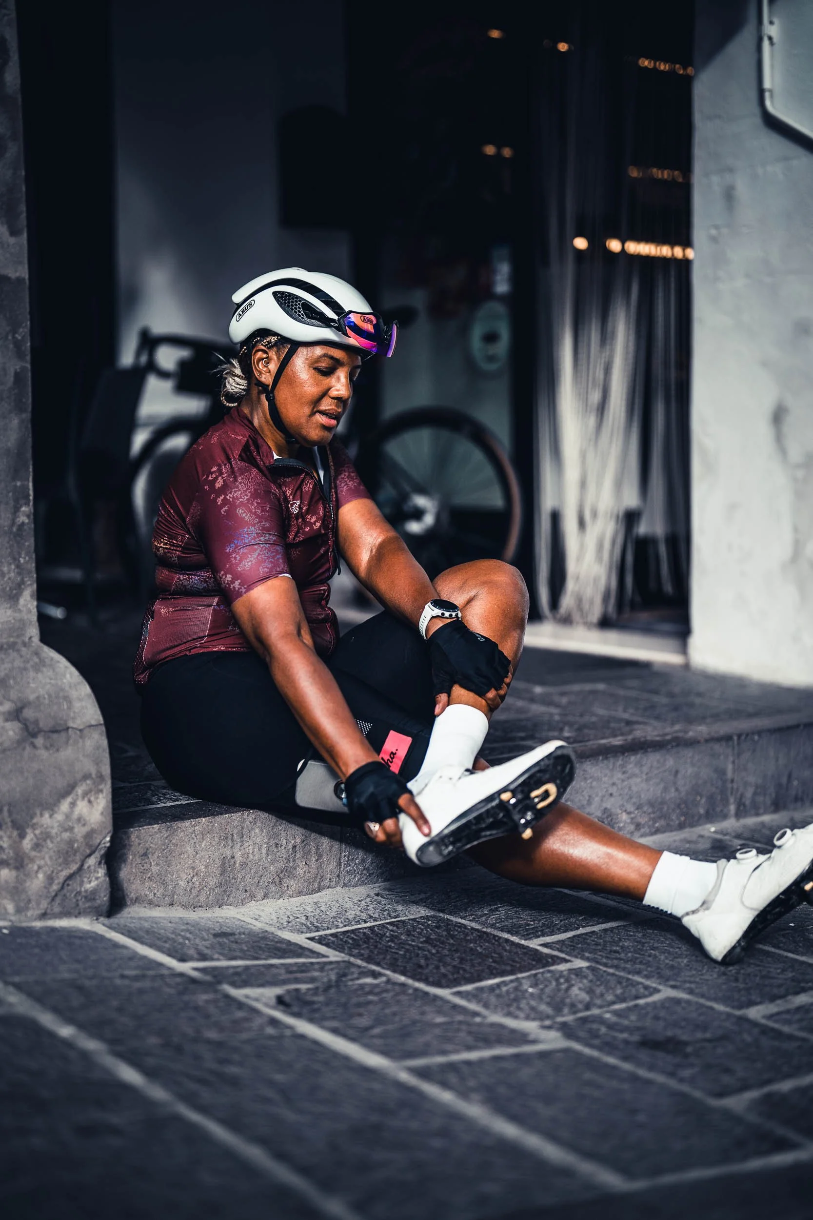 A woman in cycling gear, including a helmet and gloves, sitting on a sidewalk while putting on cycling shoes.