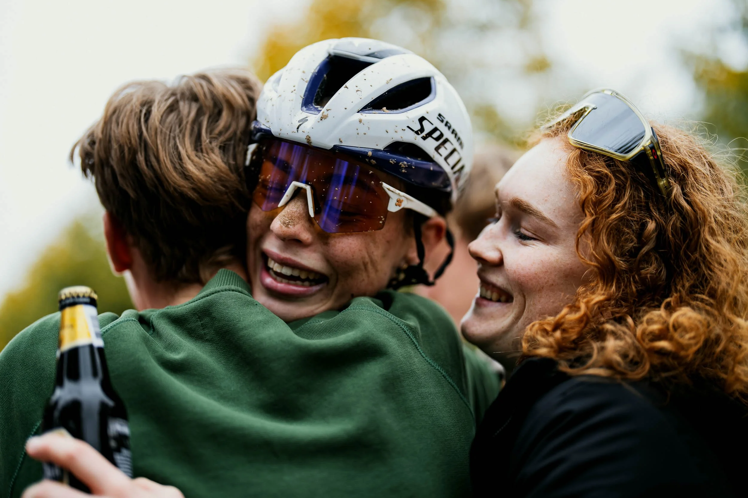 Three smiling people embracing, one wearing a helmet and sports sunglasses, holding a drink, outdoors during daytime. (Puck Pietersen) (WK Gravel)