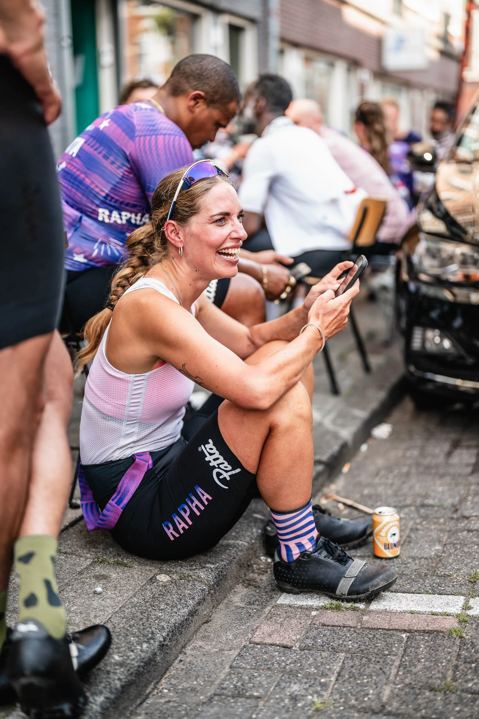 A woman sitting on a city curb, smiling and chatting on her phone, surrounded by people, wearing athletic clothes, and holding a drink can on the ground. (Patta.nl Rapha)