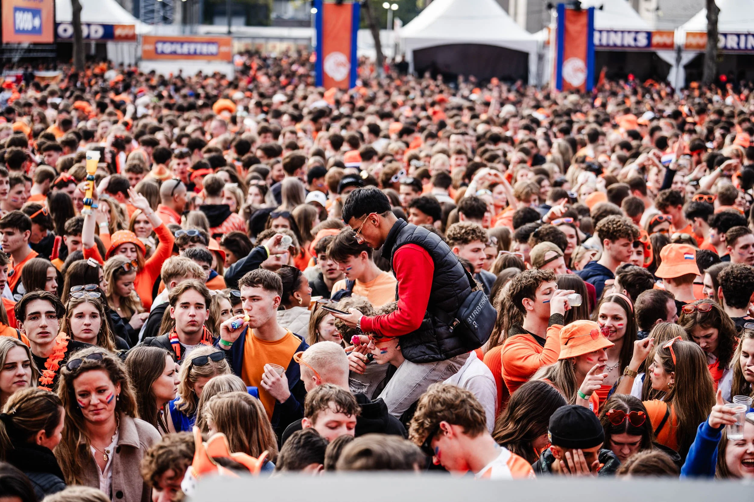 A large crowd of young people dressed in orange, gathered at an outdoor event with tents and stage in the background.