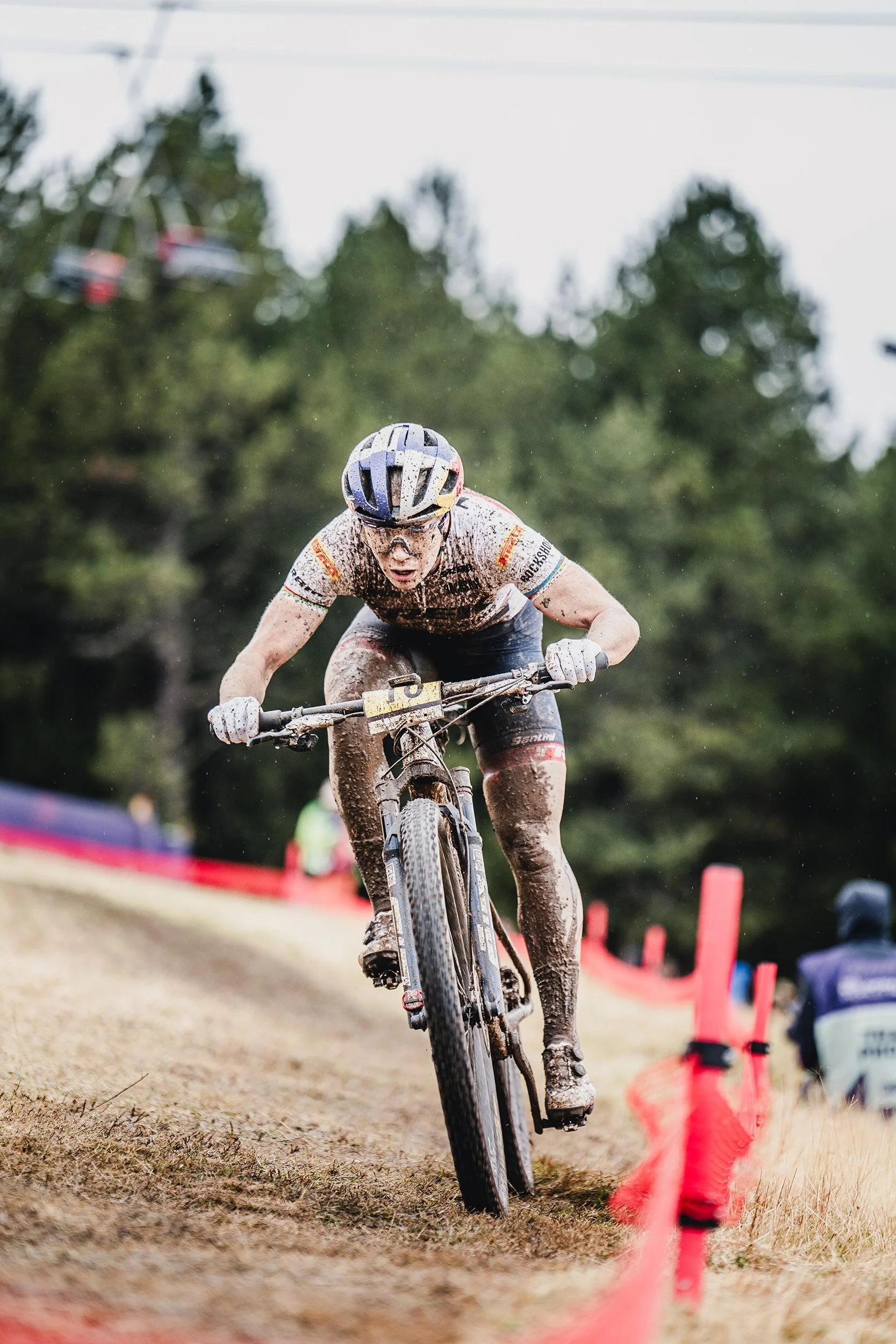 A mountain biker riding on a muddy trail, leaning forward with focus, wearing a helmet, goggles, and muddy cycling gear, with a red barrier fence lining the trail and trees in the background.