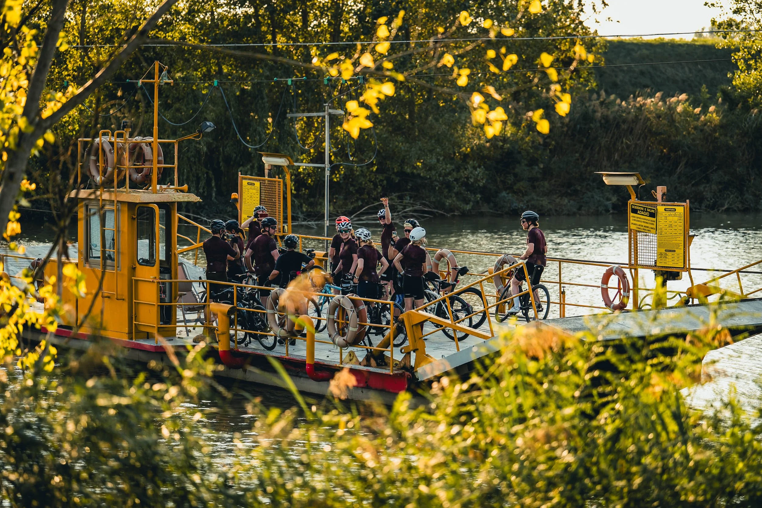 A group of cyclists gathering on a yellow ferryboat on a river, with boats and trees in the background, during late afternoon or early evening.