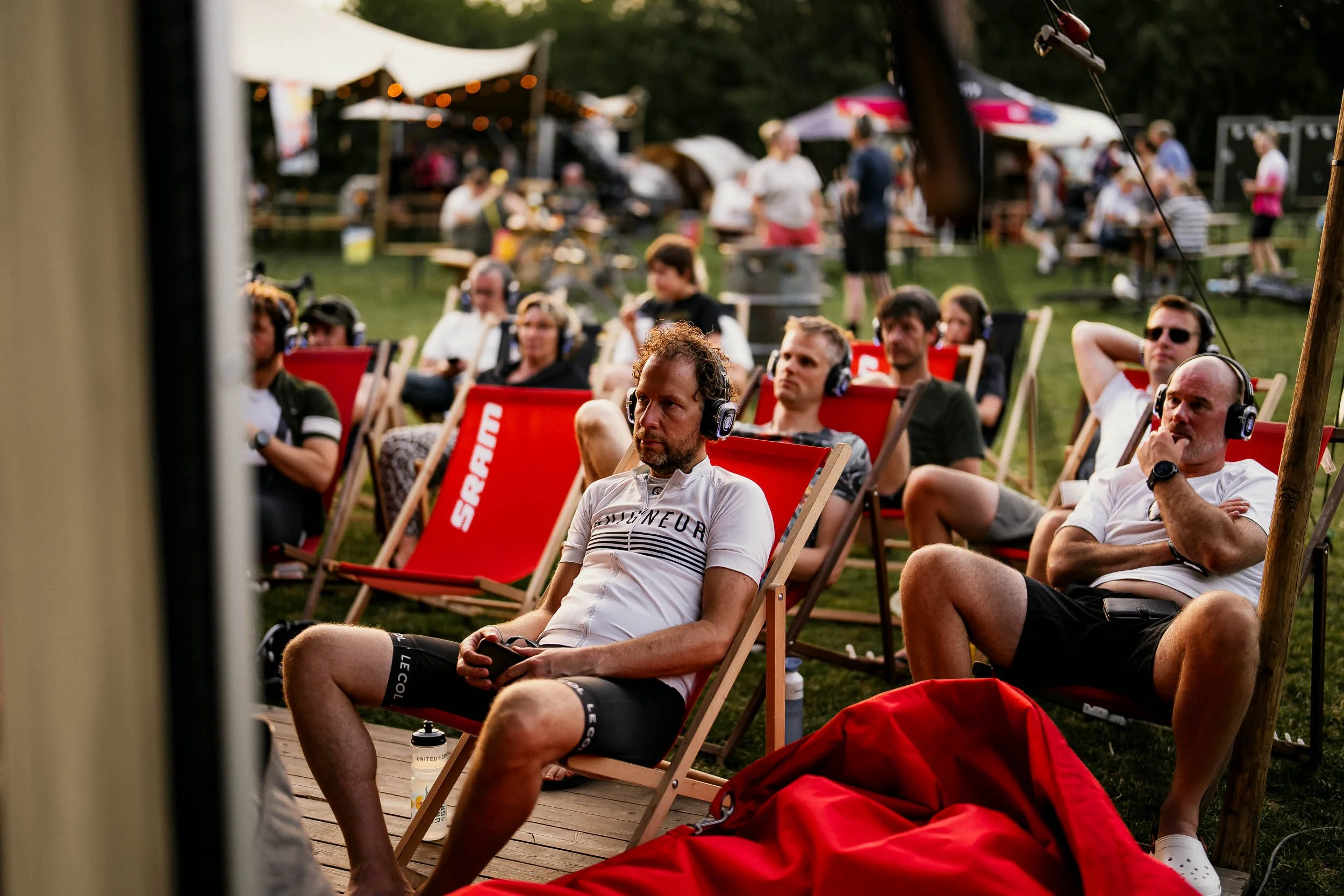 People sitting outdoors on deck chairs, listening to music with headphones at a festival or outdoor event during the evening, with tents, umbrellas, and other attendees in the background.