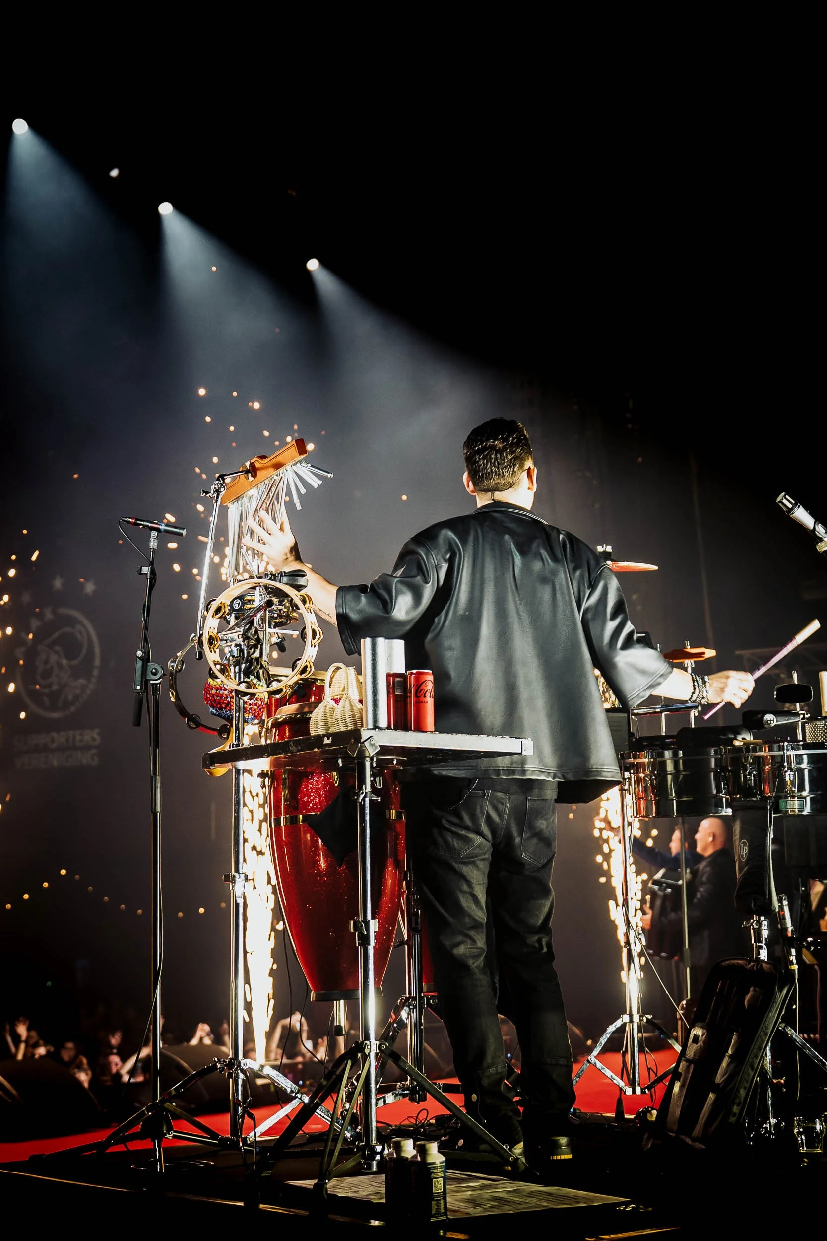 Performer playing percussion instruments on stage during a concert, with sparks and lights in the background.